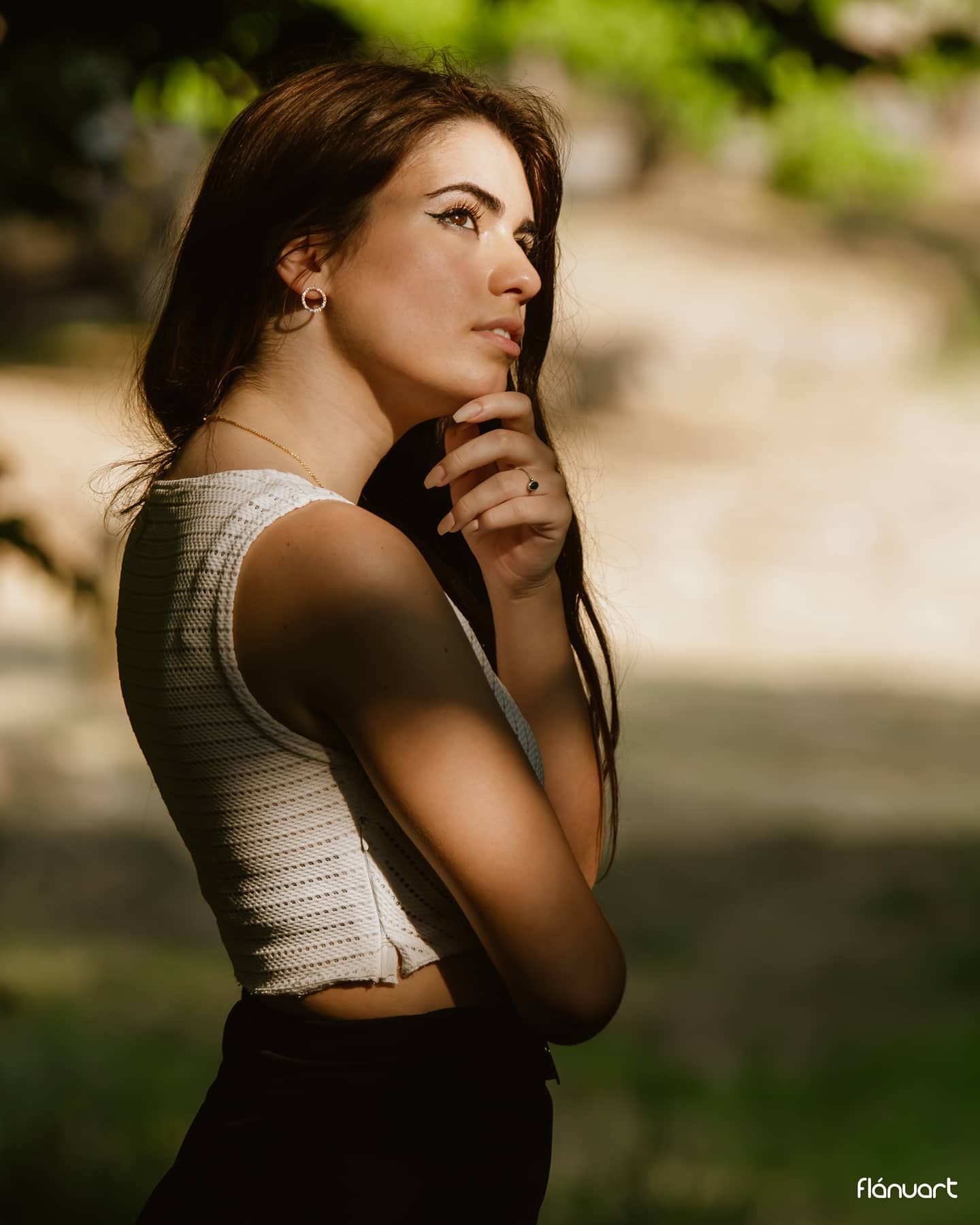 Brunette woman in white knit crop top posing thoughtfully in dappled sunlight outdoor setting.