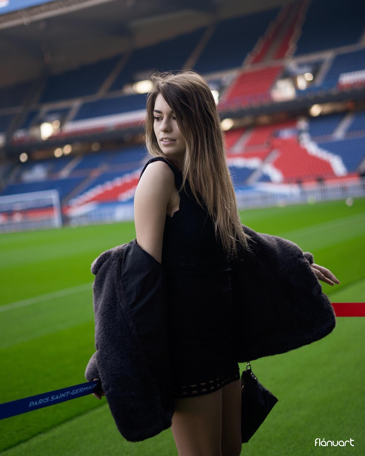 Young woman in black dress holding fur coat, posing on football pitch inside stadium.