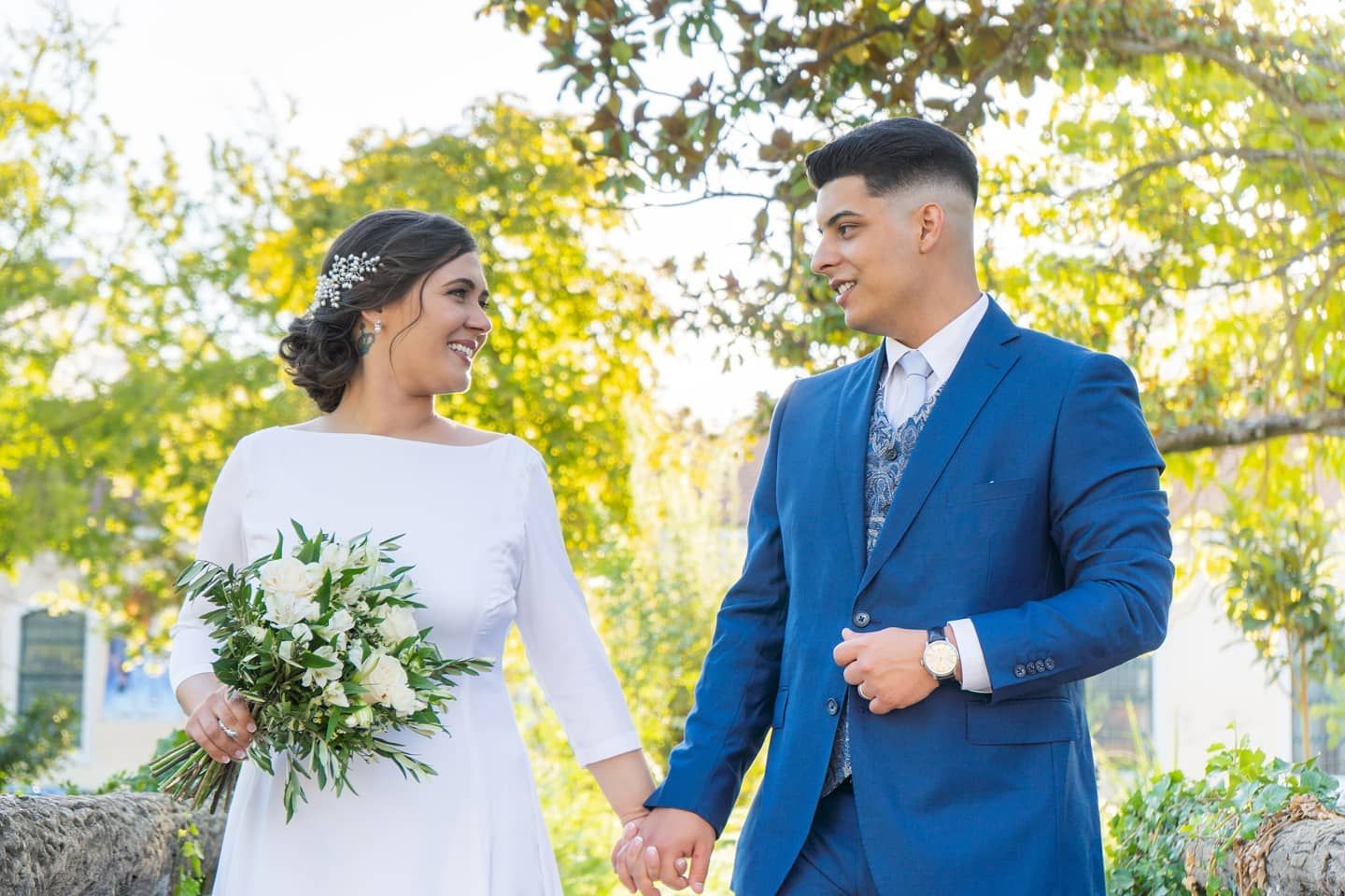 Smiling bride holding white bouquet walks hand in hand with groom in blue suit outdoors.