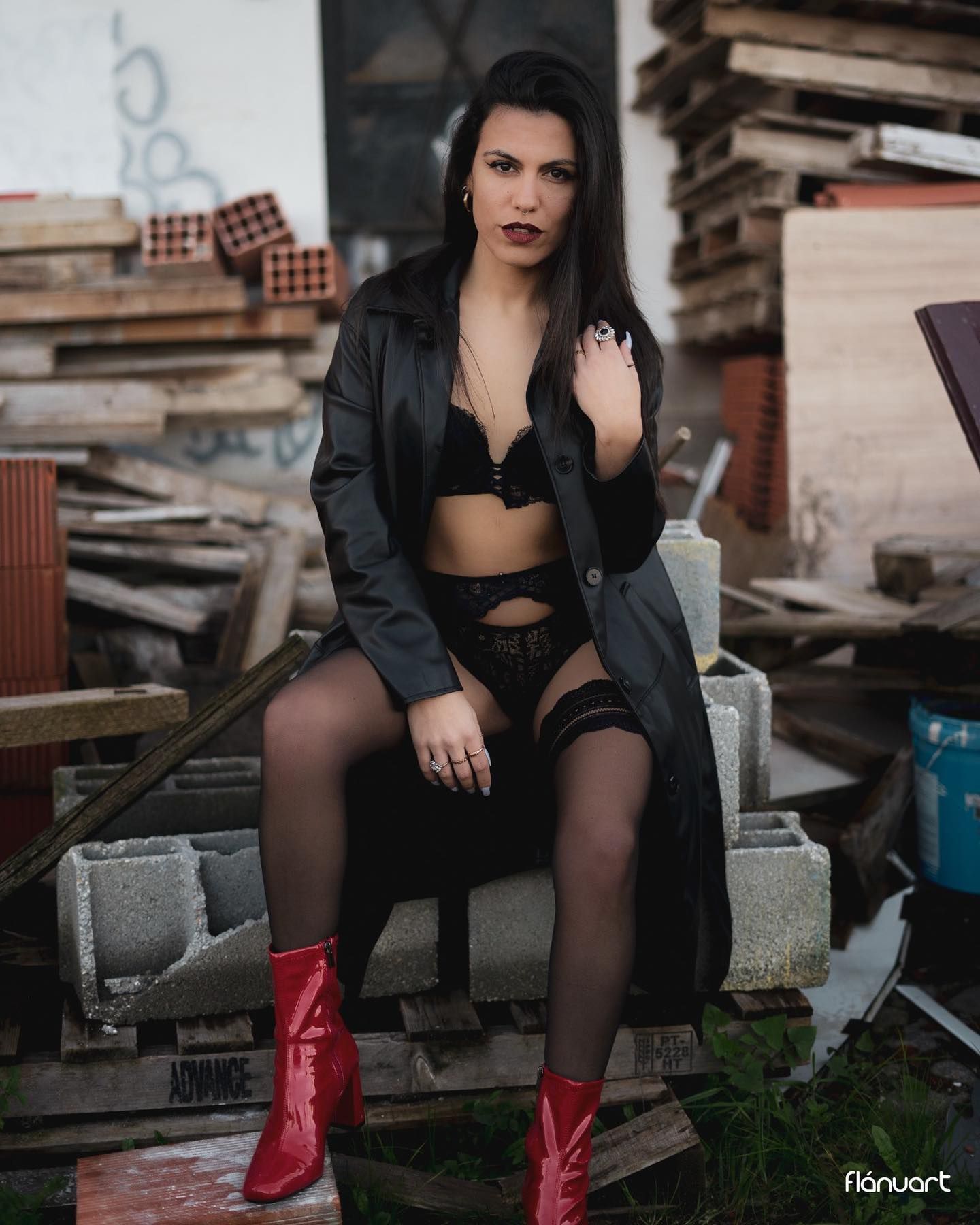 Woman in black lingerie and leather coat sitting in industrial ruins wearing red boots.