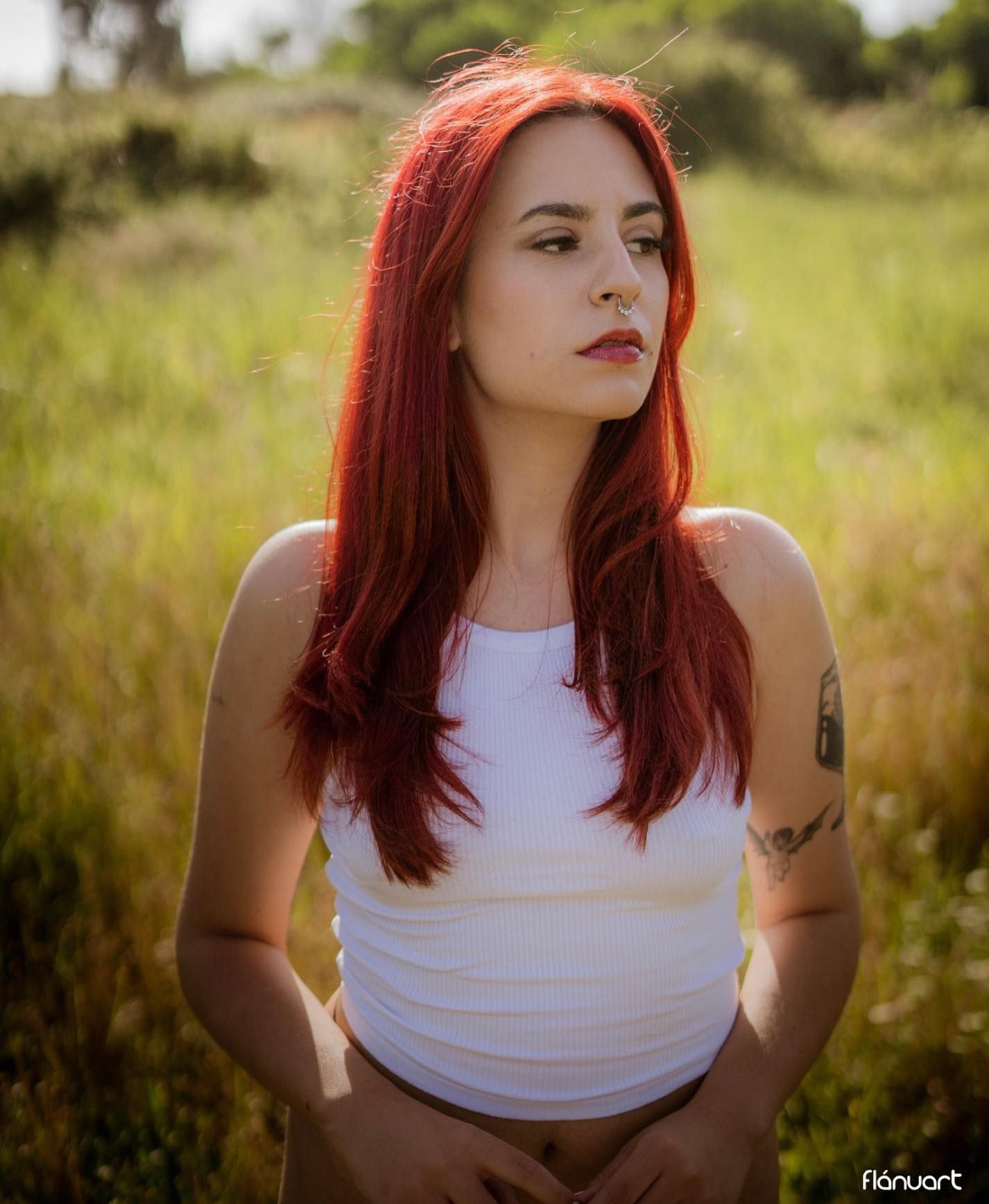 Young woman with vibrant red hair wearing white crop top posing in sunny green field.
