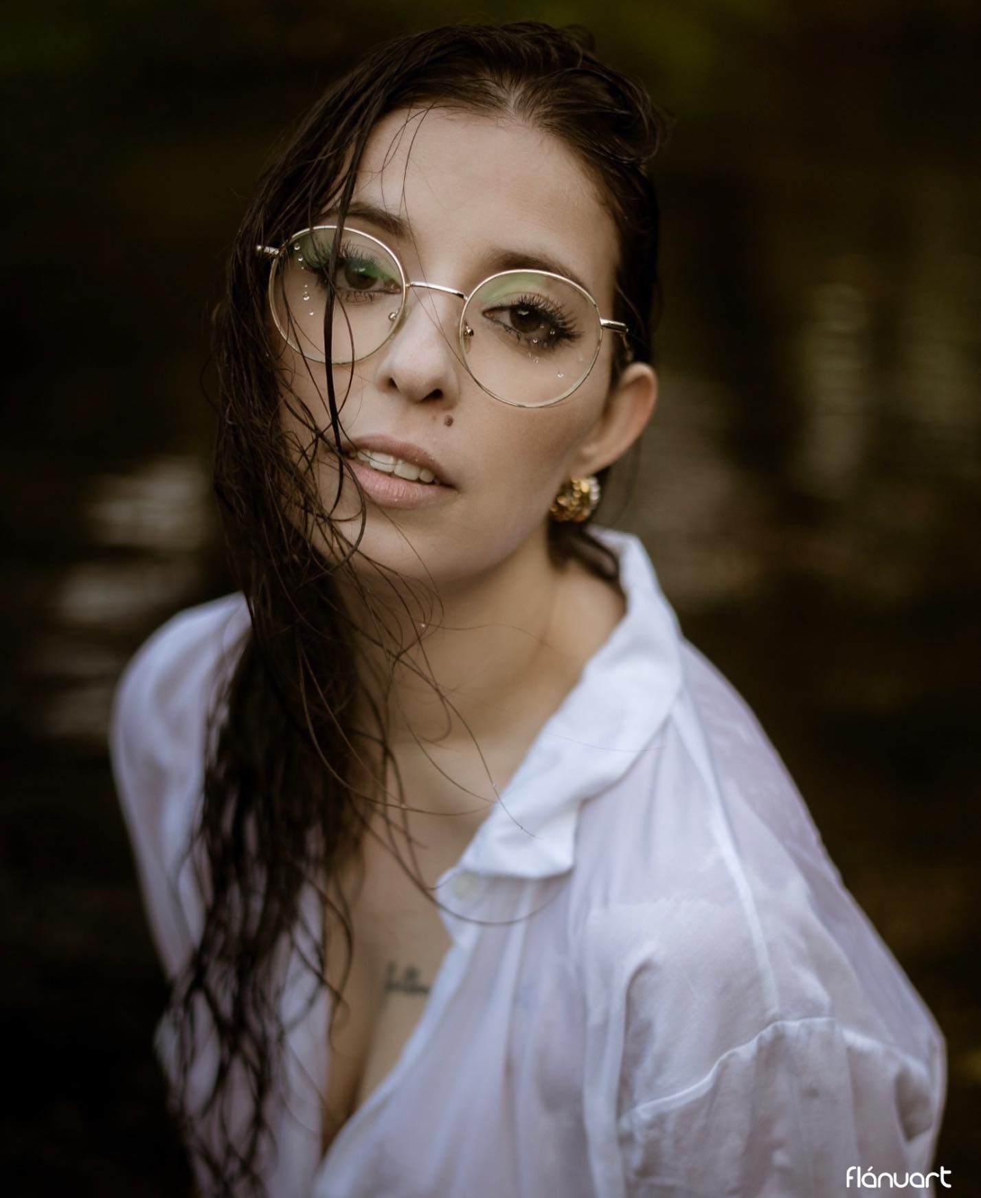 Woman with wet dark hair and round glasses wearing white shirt in moody outdoor water setting.