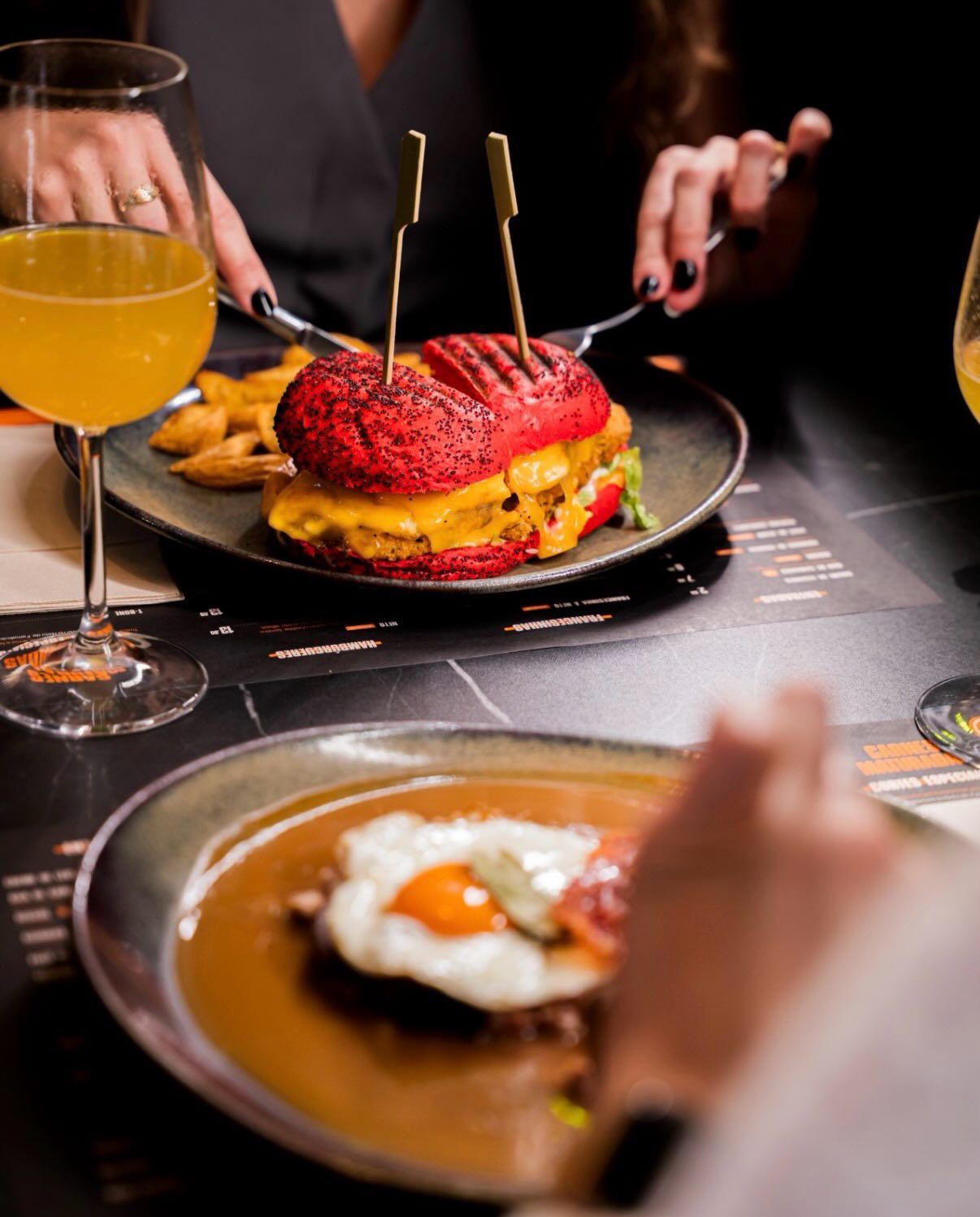 A vibrant red burger with gold picks served on a dark plate alongside a fried egg dish and orange cocktails.
