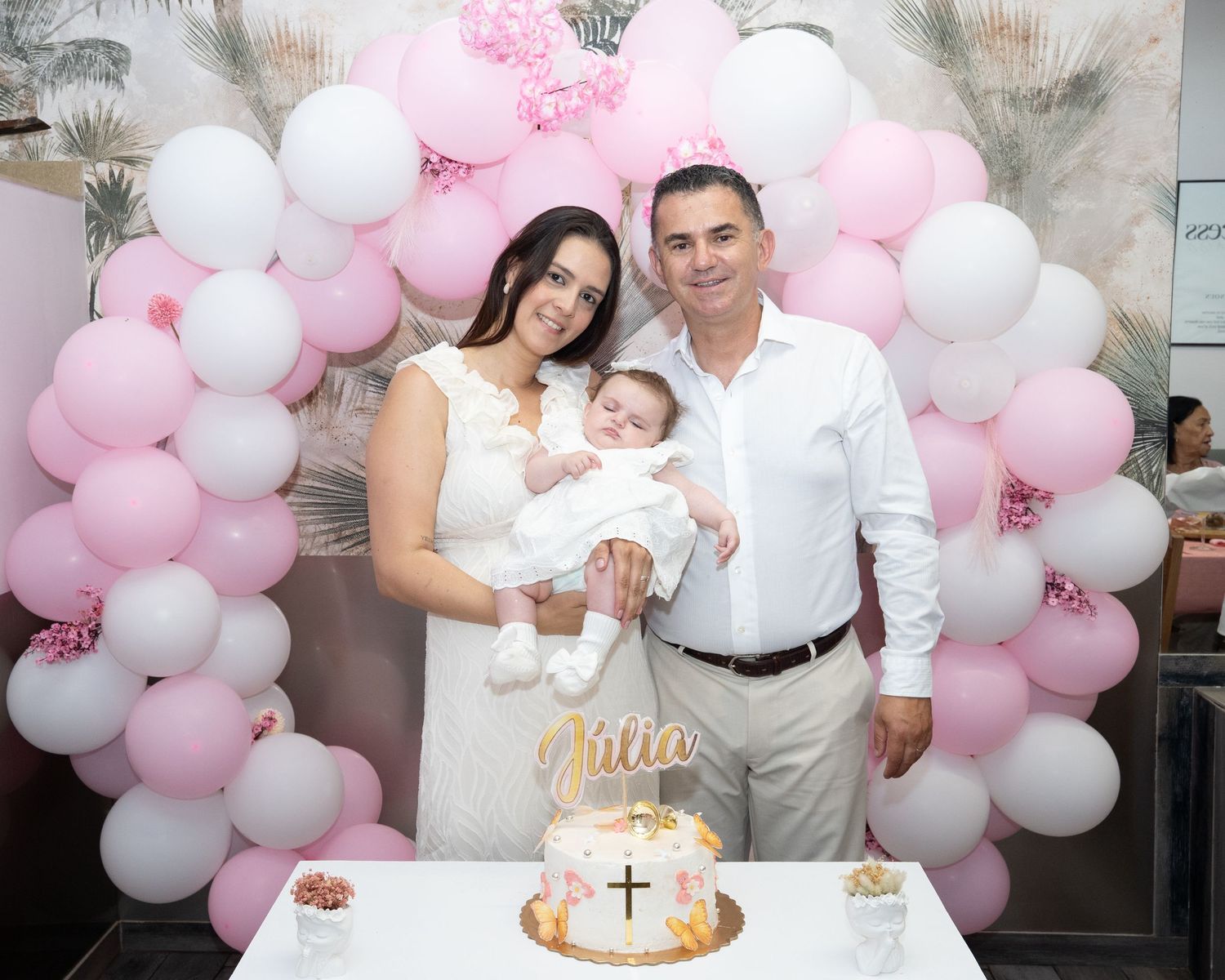 Family celebrating a baby girl baptism with pink balloon arch and decorated cross cake.