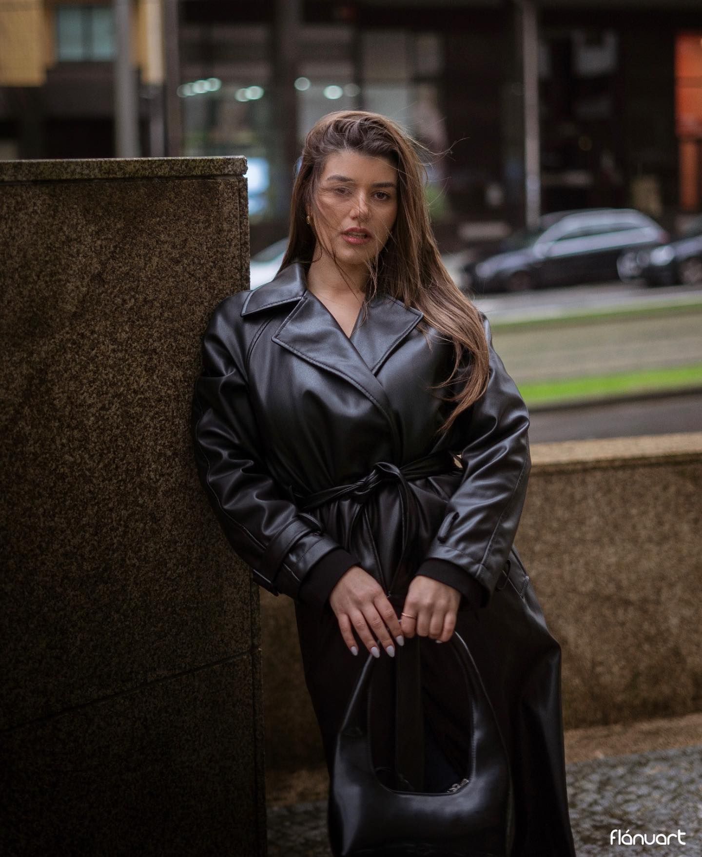 Young woman in black leather trench coat posing outdoors on urban street with blurred city background.