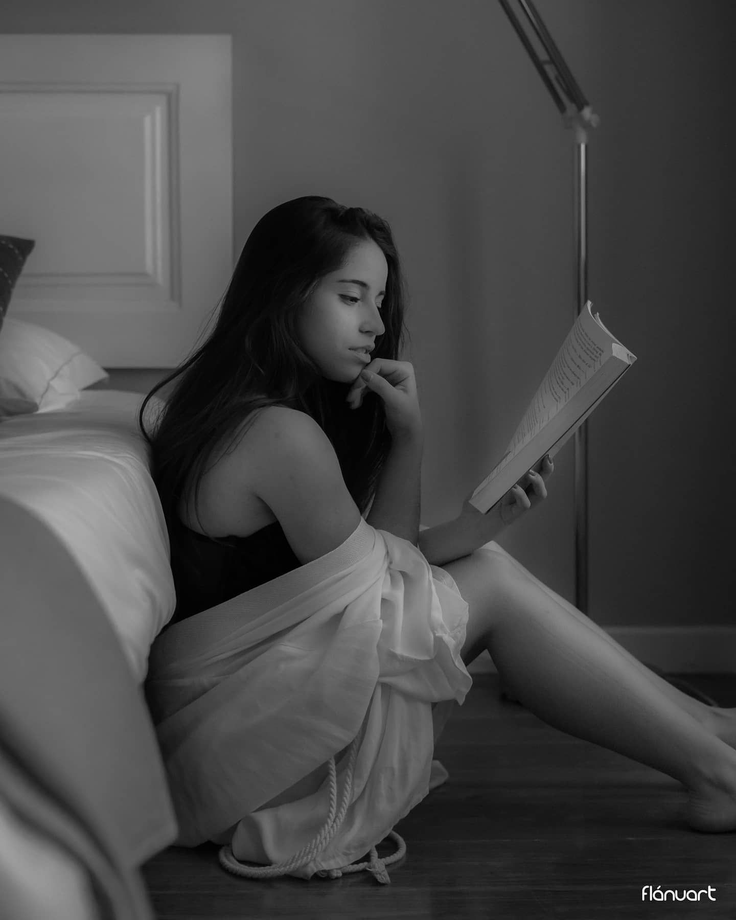 Black and white photo of a woman sitting on the floor reading a book by a bed.