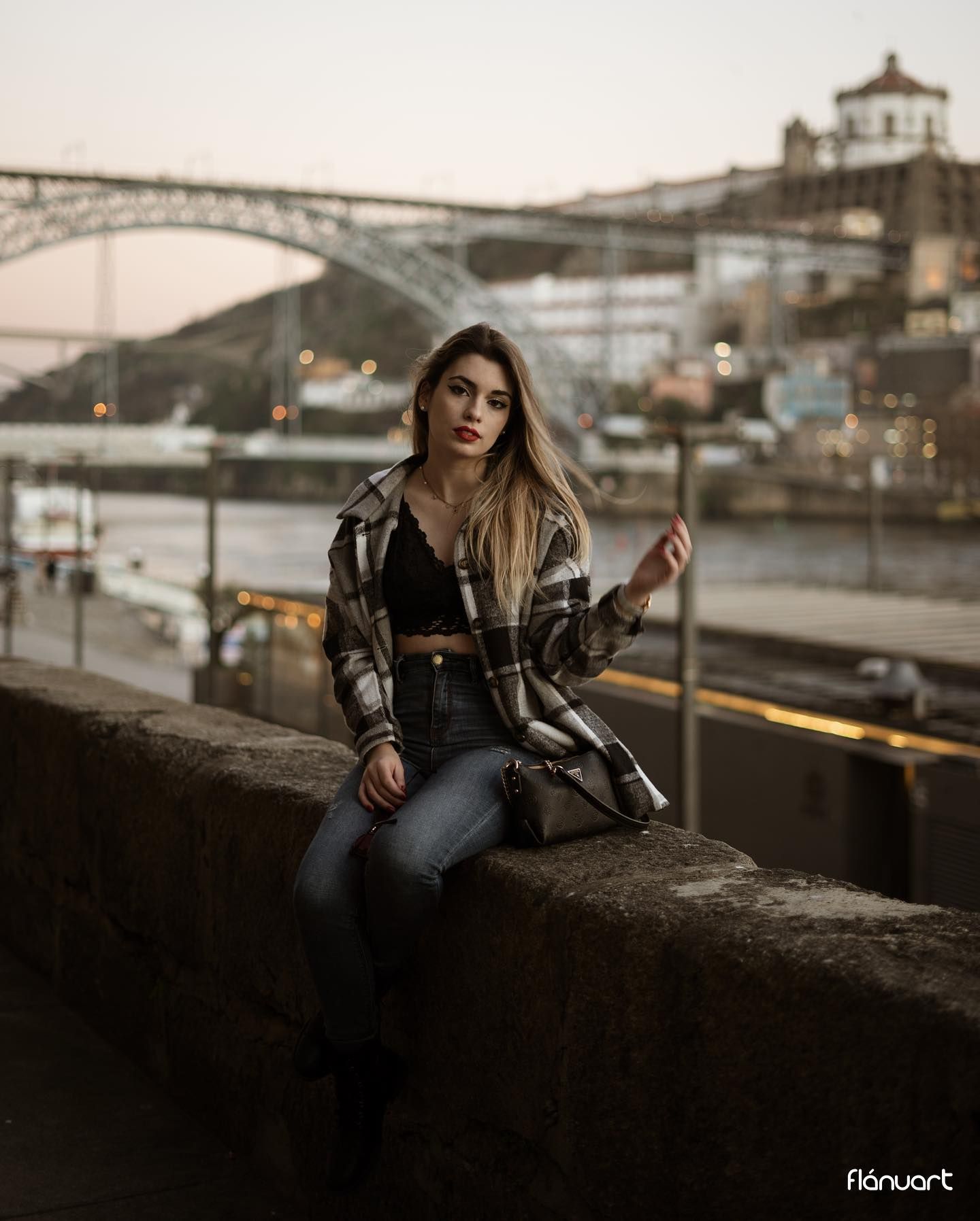 Young woman in plaid shirt, black crop top, and jeans sitting on a wall with a bridge and river at dusk.