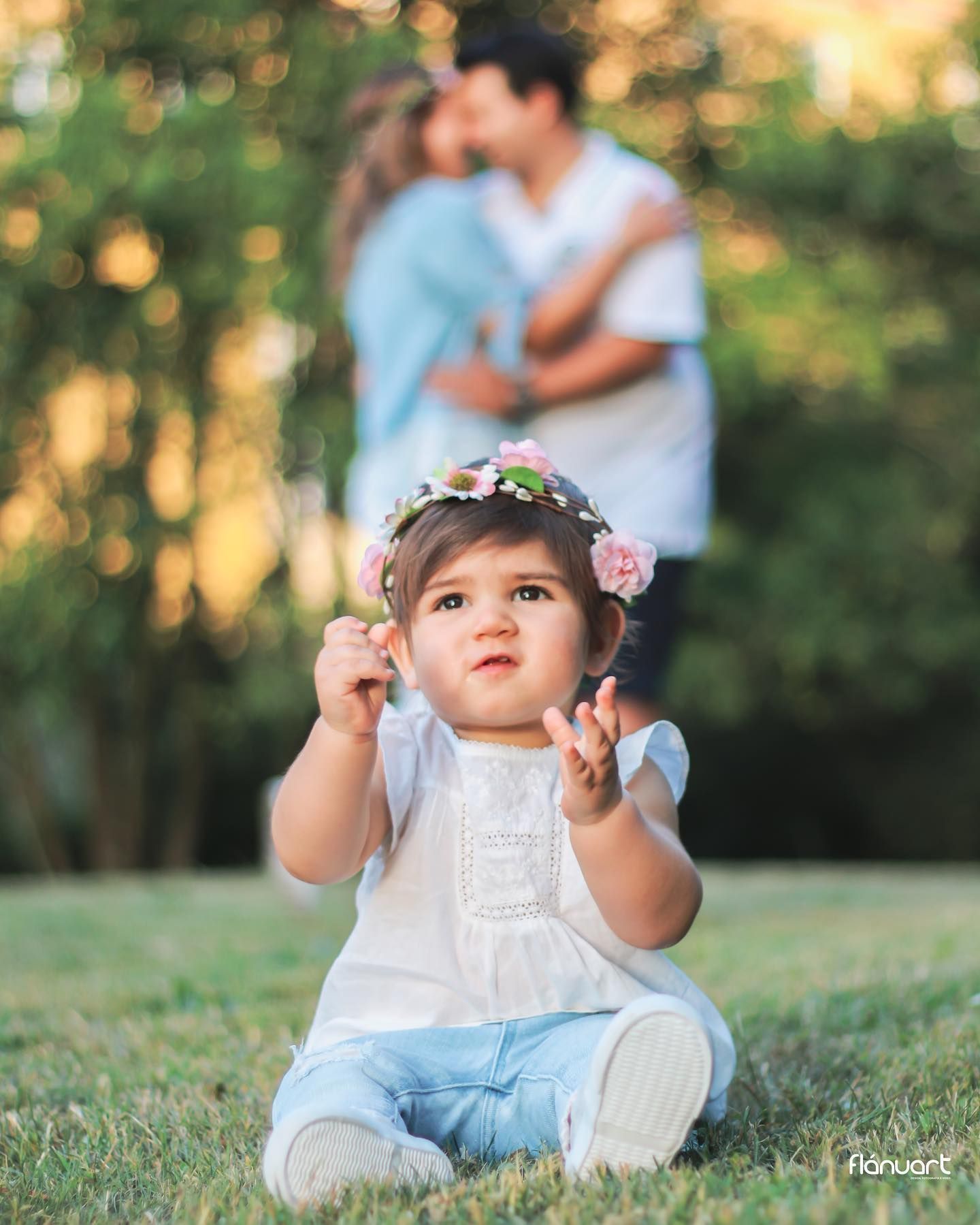 Adorable baby girl with flower crown sitting on grass while parents share a kiss behind her.