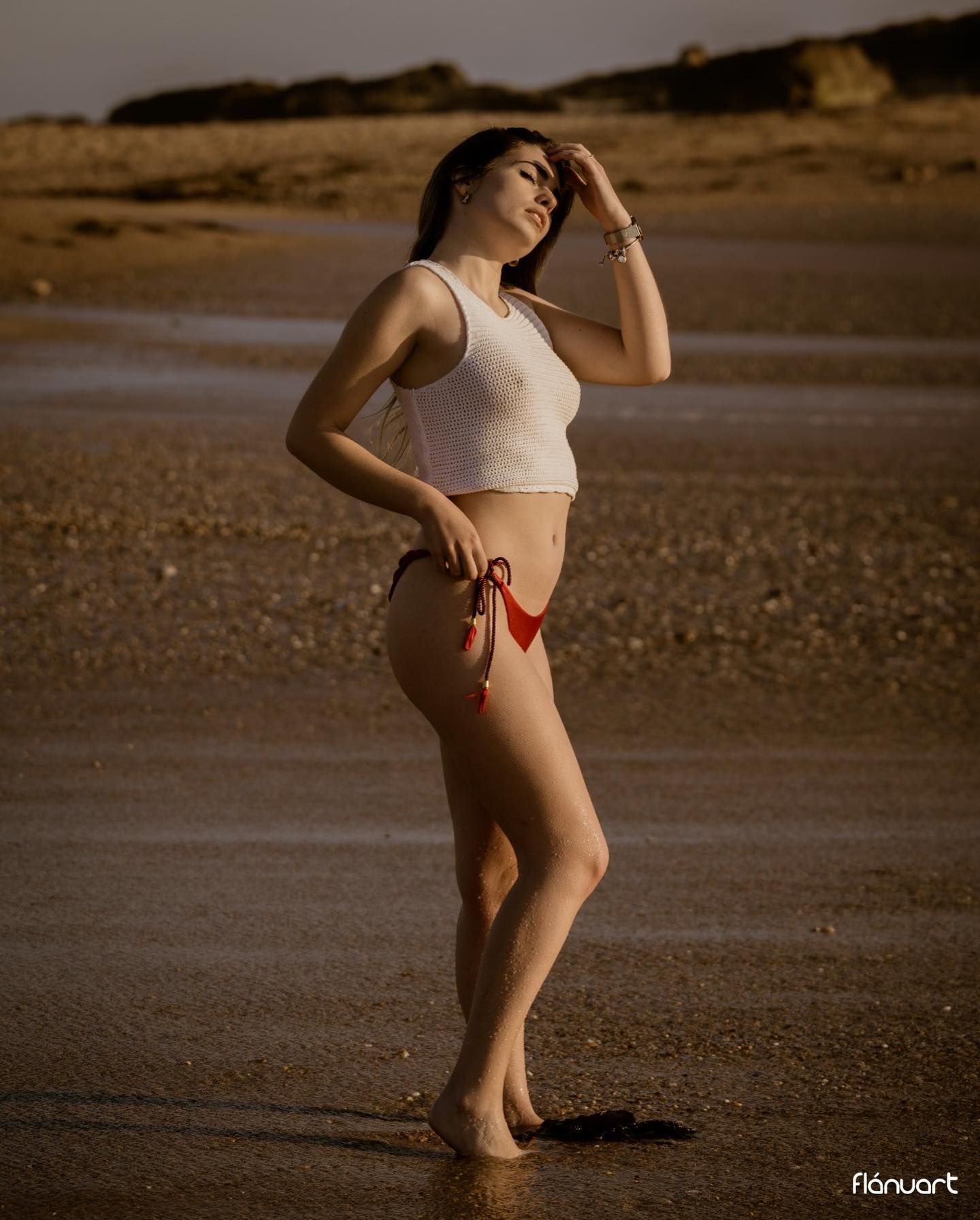 Brunette woman in white tank top and red bikini bottom posing on wet beach.