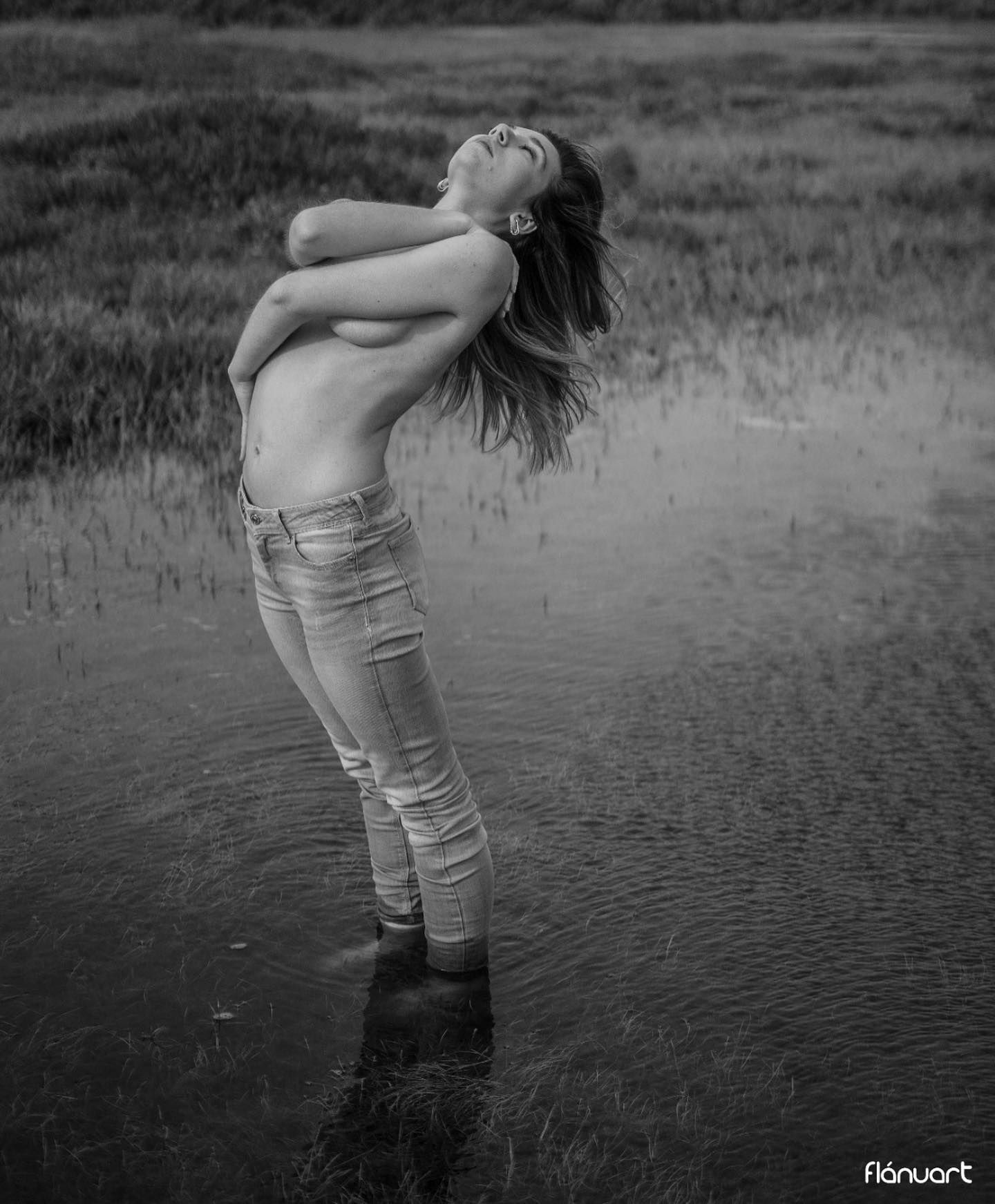 Black and white photo of a topless woman in jeans standing in water arching back.