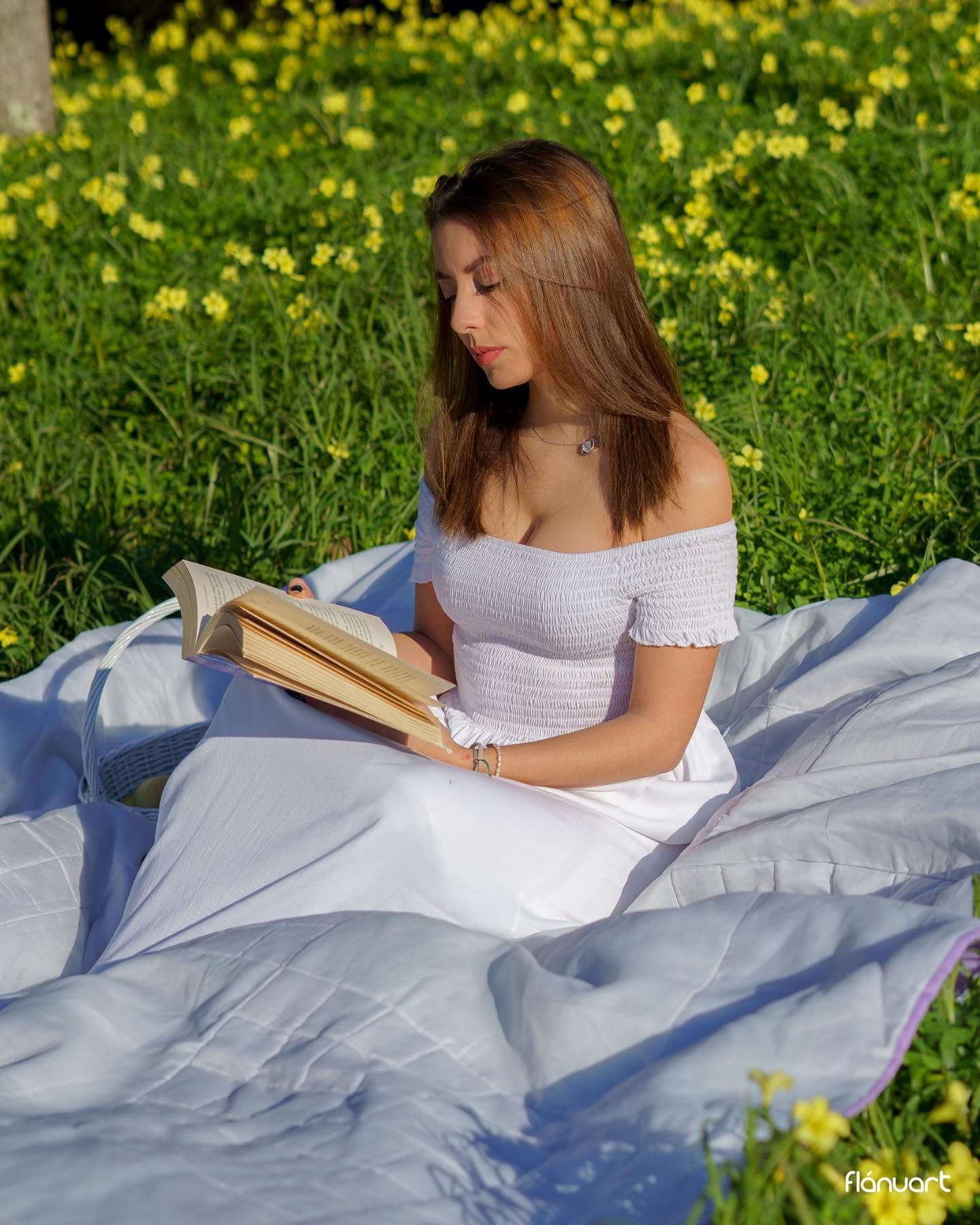 Young woman in white off-shoulder dress reads a book while sitting on a blanket in a sunny field of yellow flowers.