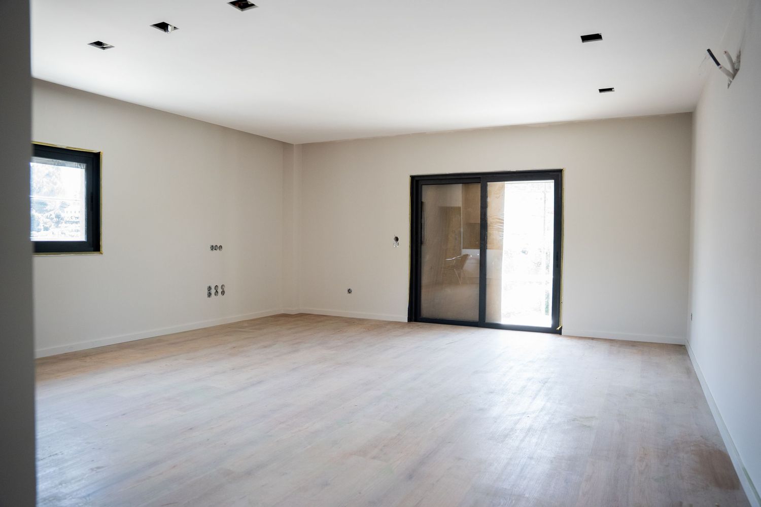 Empty modern room with light wood floors, recessed lighting, white walls, a small window, and sliding glass door.