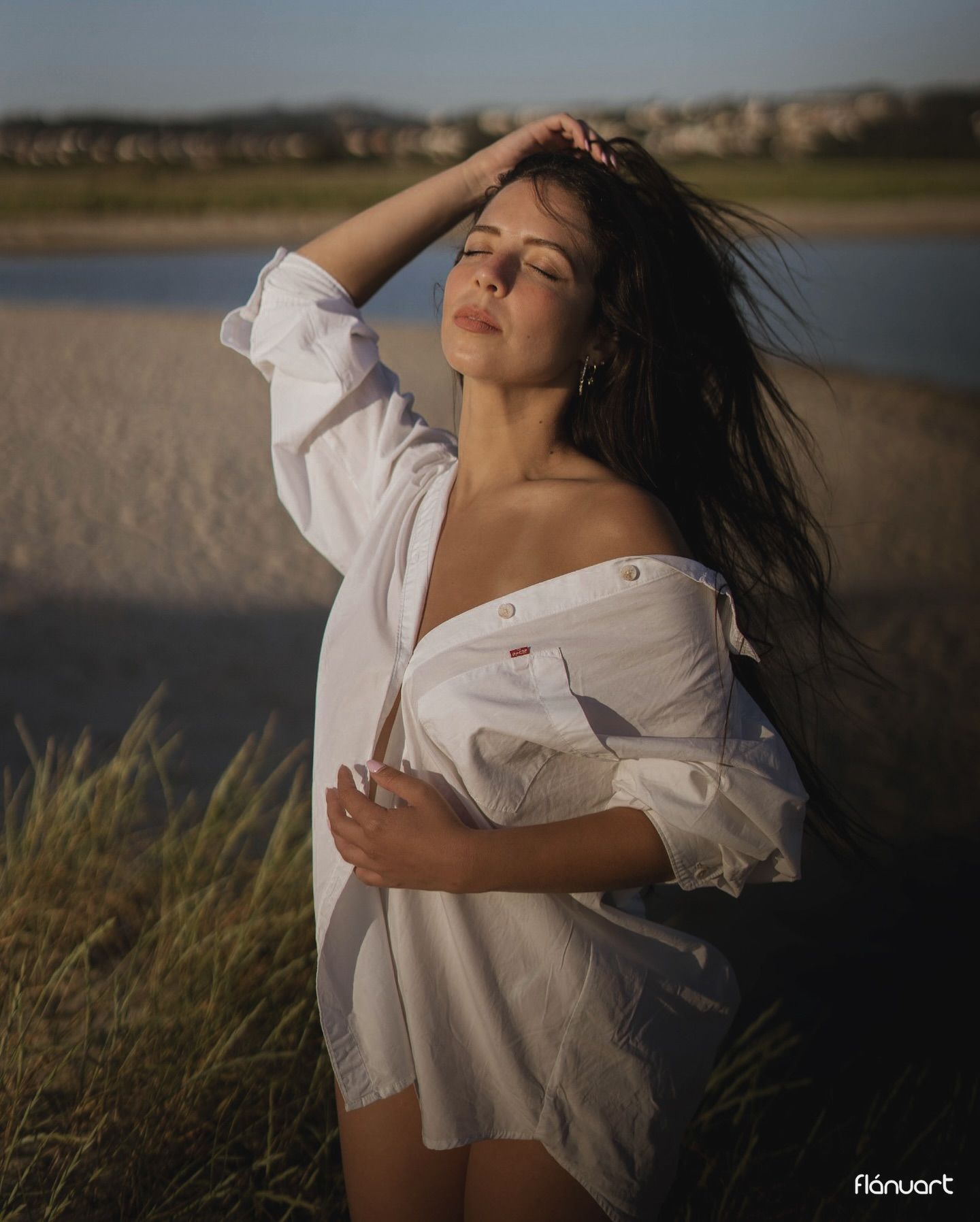 Woman in open white shirt posing outdoors near sandy shore at dusk.