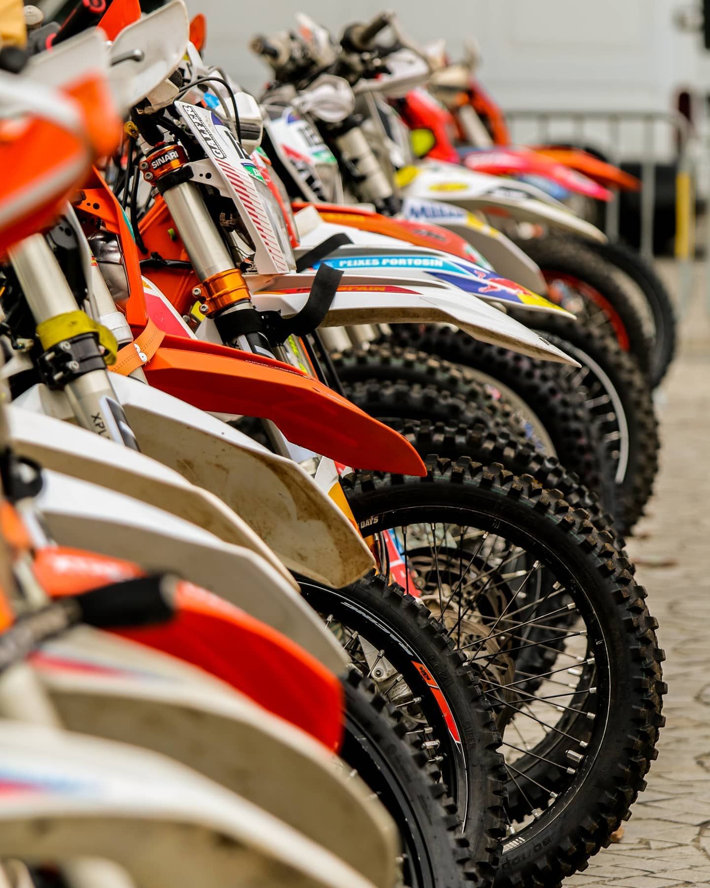 Row of colorful KTM dirt bikes lined up at a motocross racing event on a paved surface.