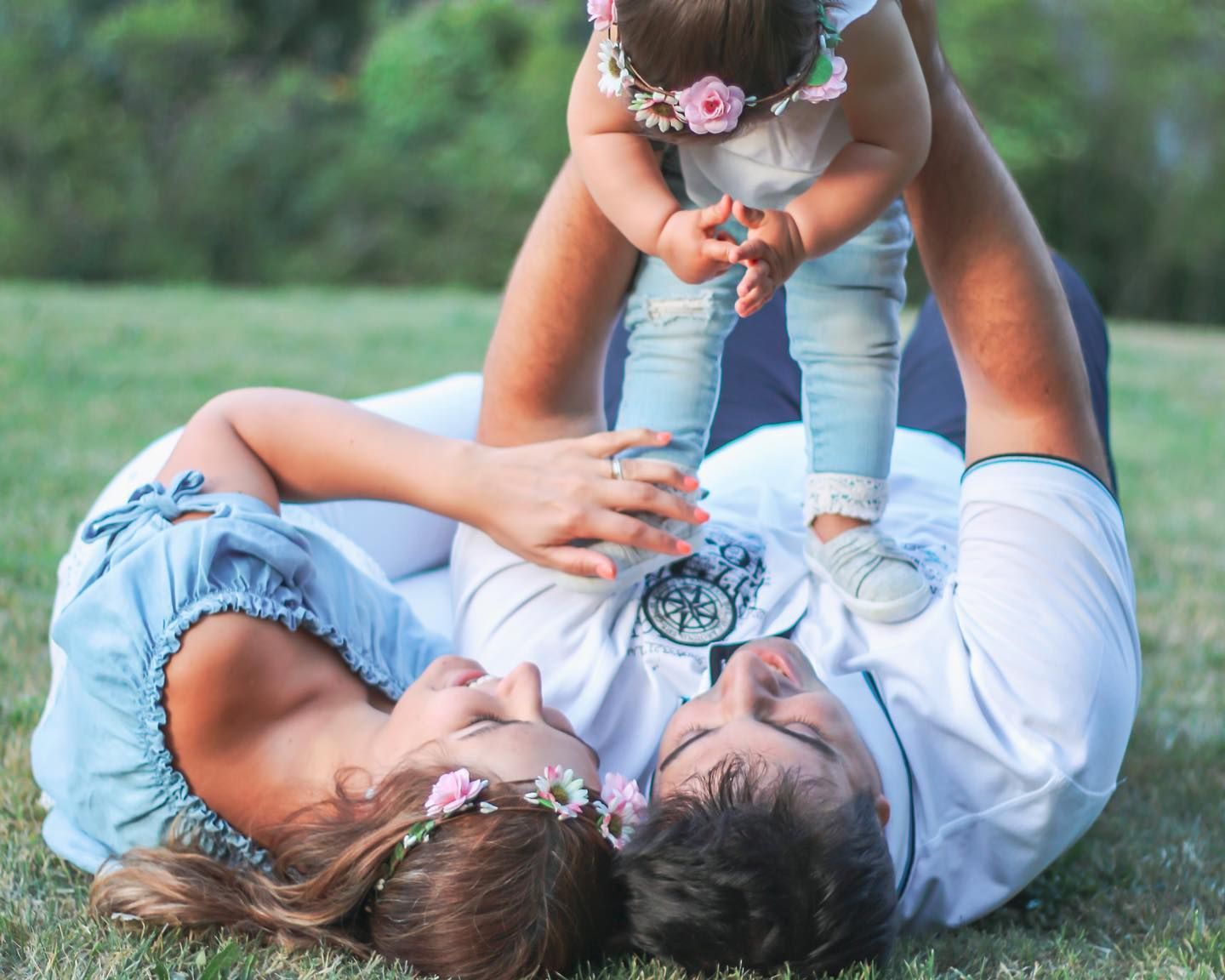 Family playing on grass, parents lifting baby girl wearing floral crown in joyful outdoor moment.