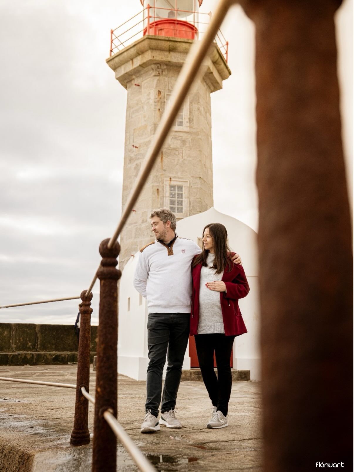 Couple walking together near historic lighthouse on overcast day, woman in red jacket.