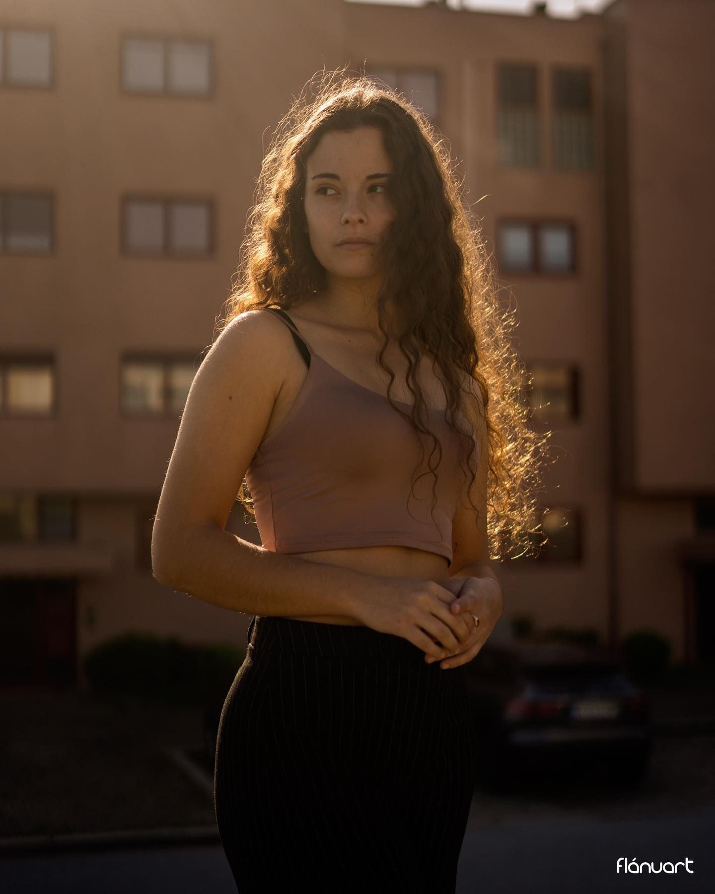 Young woman with curly brown hair in a beige crop top and black skirt, posing at golden hour outdoors.