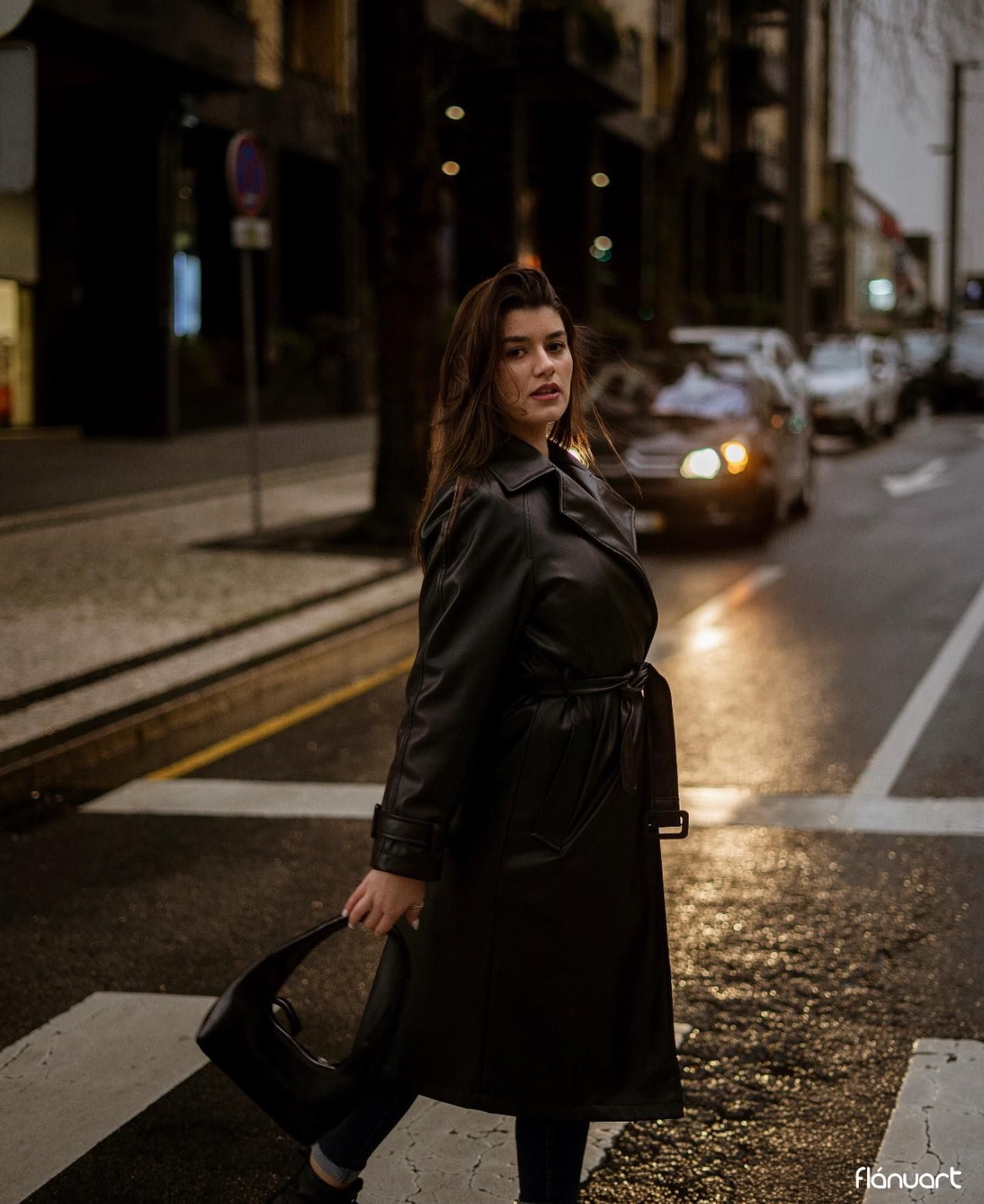 Woman in black leather coat crossing illuminated city street at night.