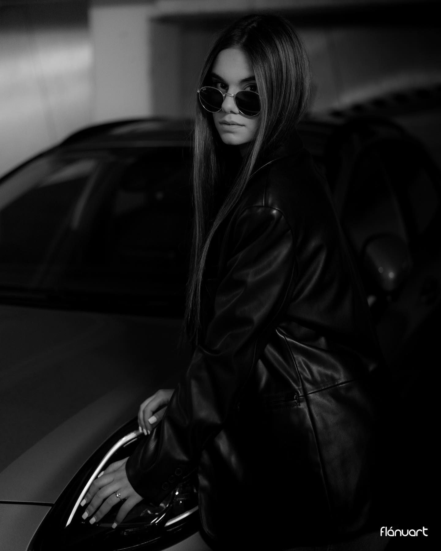 Black and white portrait of a woman in leather coat and round sunglasses leaning on a car.