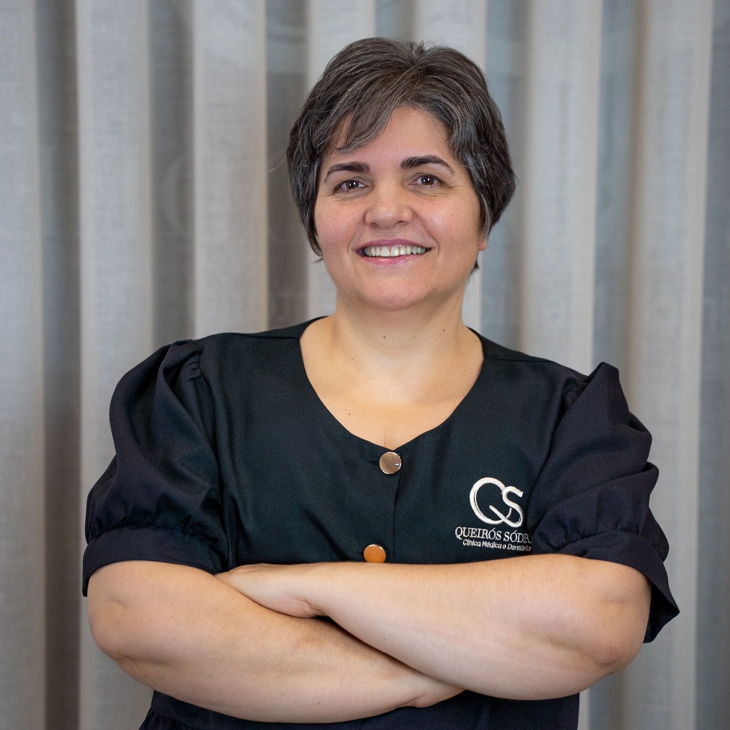 Middle-aged female healthcare worker in black scrubs smiling with arms crossed in a professional portrait.