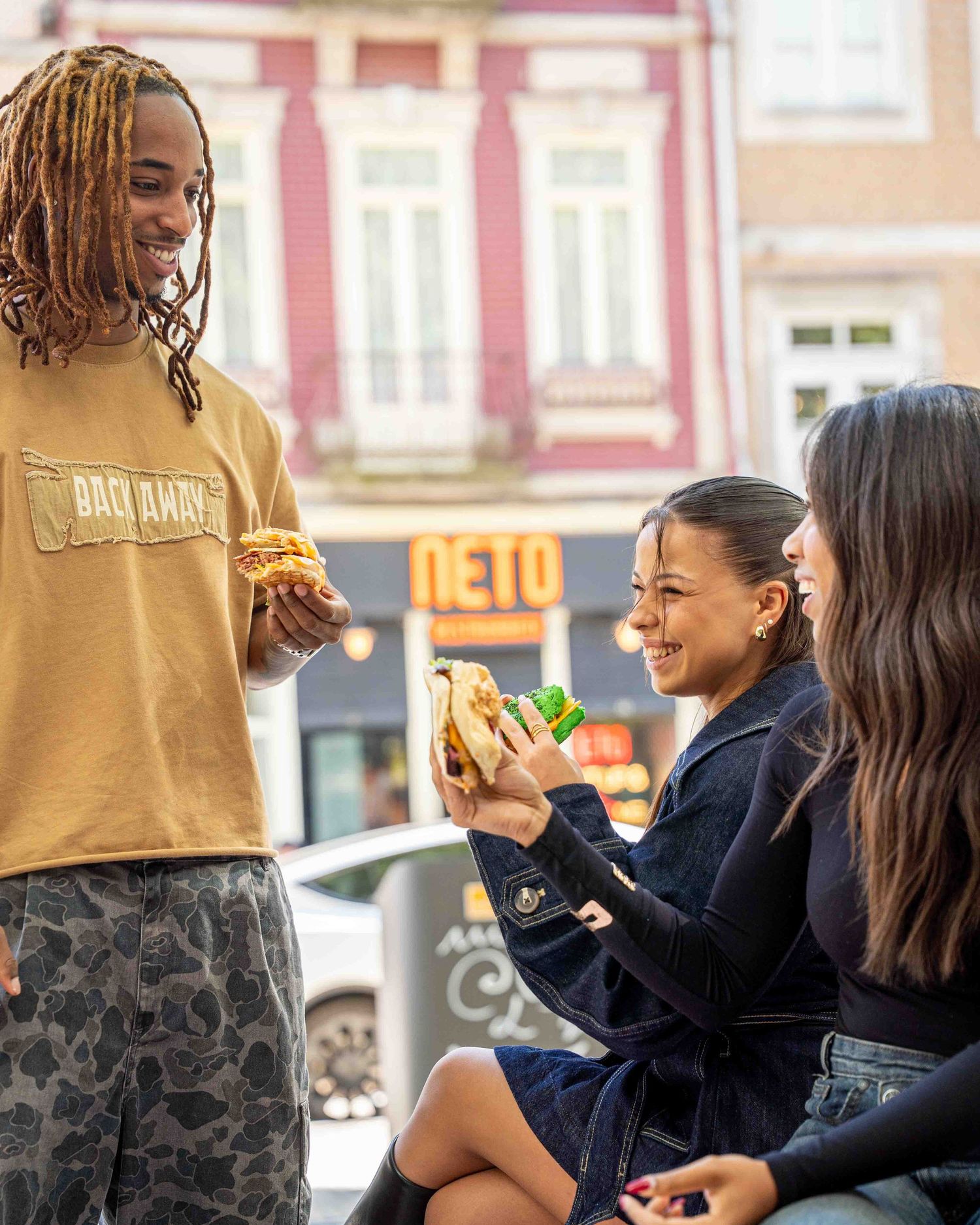 Three friends enjoying sandwiches outdoors near Neto Restaurante on a sunny day in the city.