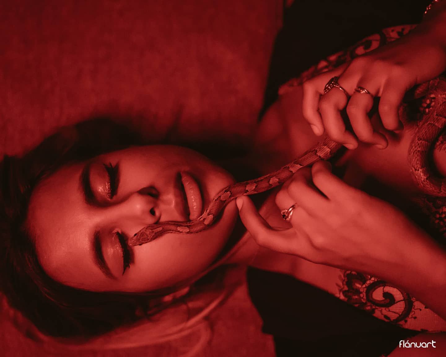 Woman lying back with a snake near her face in dramatic red-toned artistic photography.