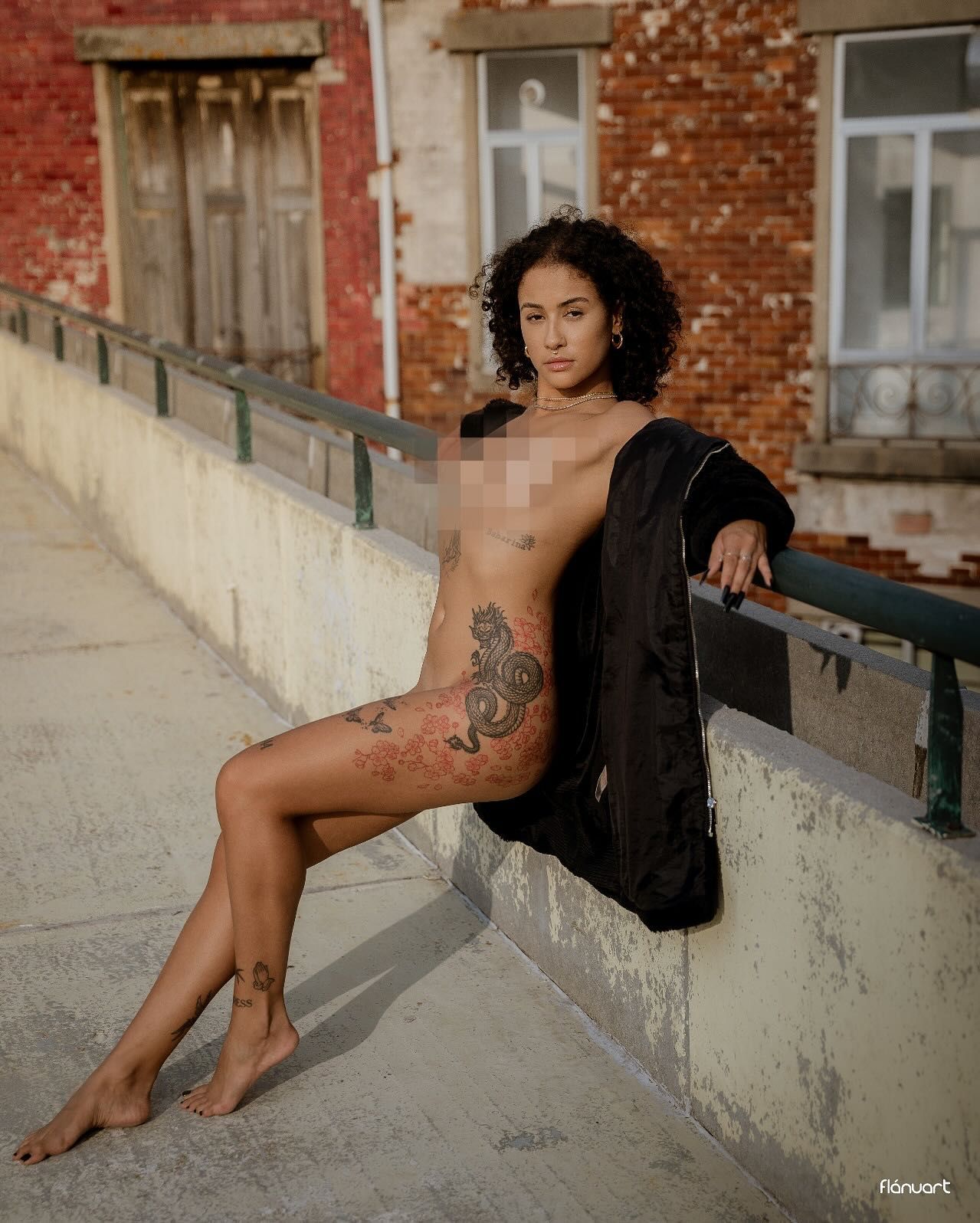 Tattooed woman posing outdoors on a balcony beside a rustic brick building.