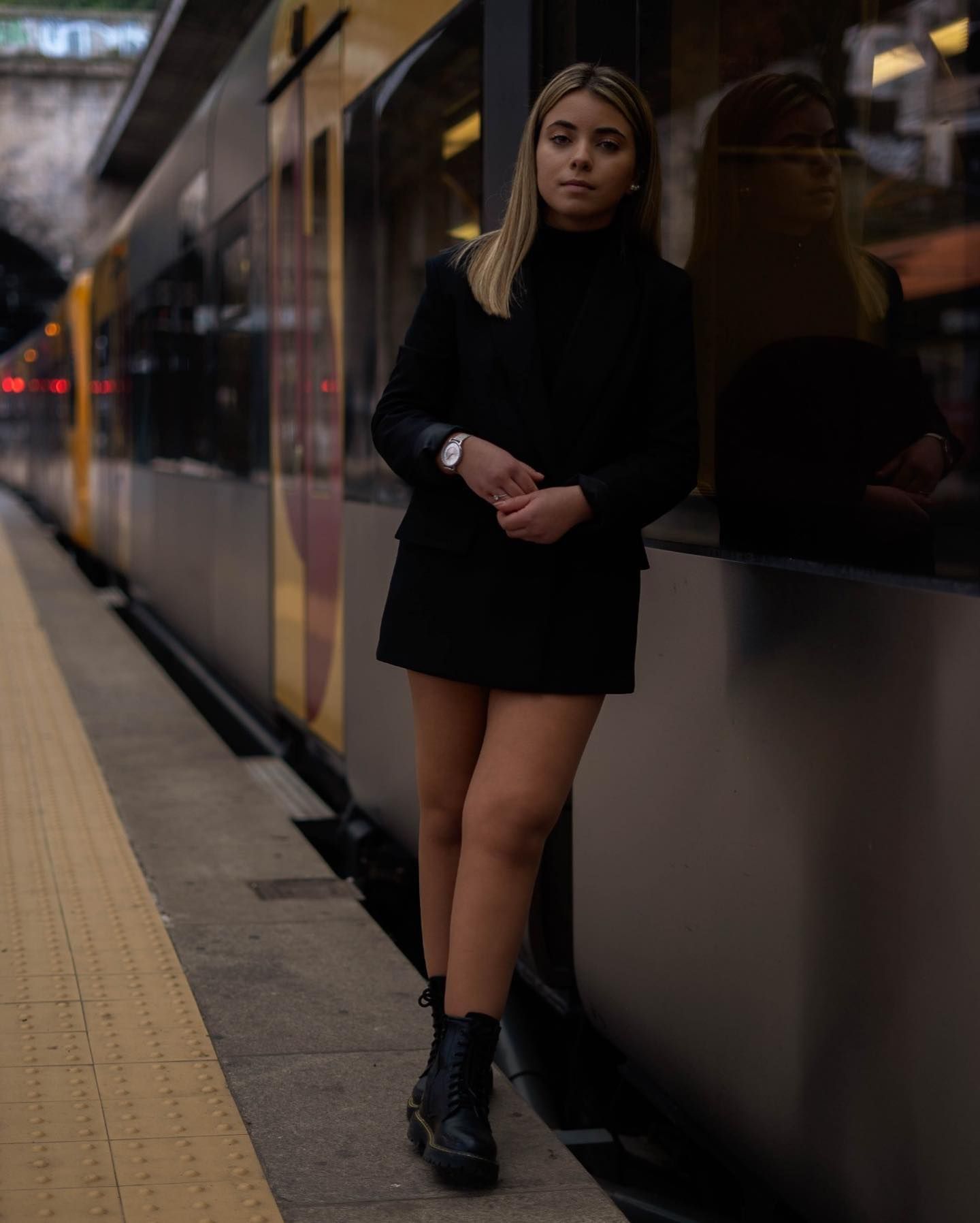 Blonde woman in black blazer and mini skirt standing beside train at station platform.