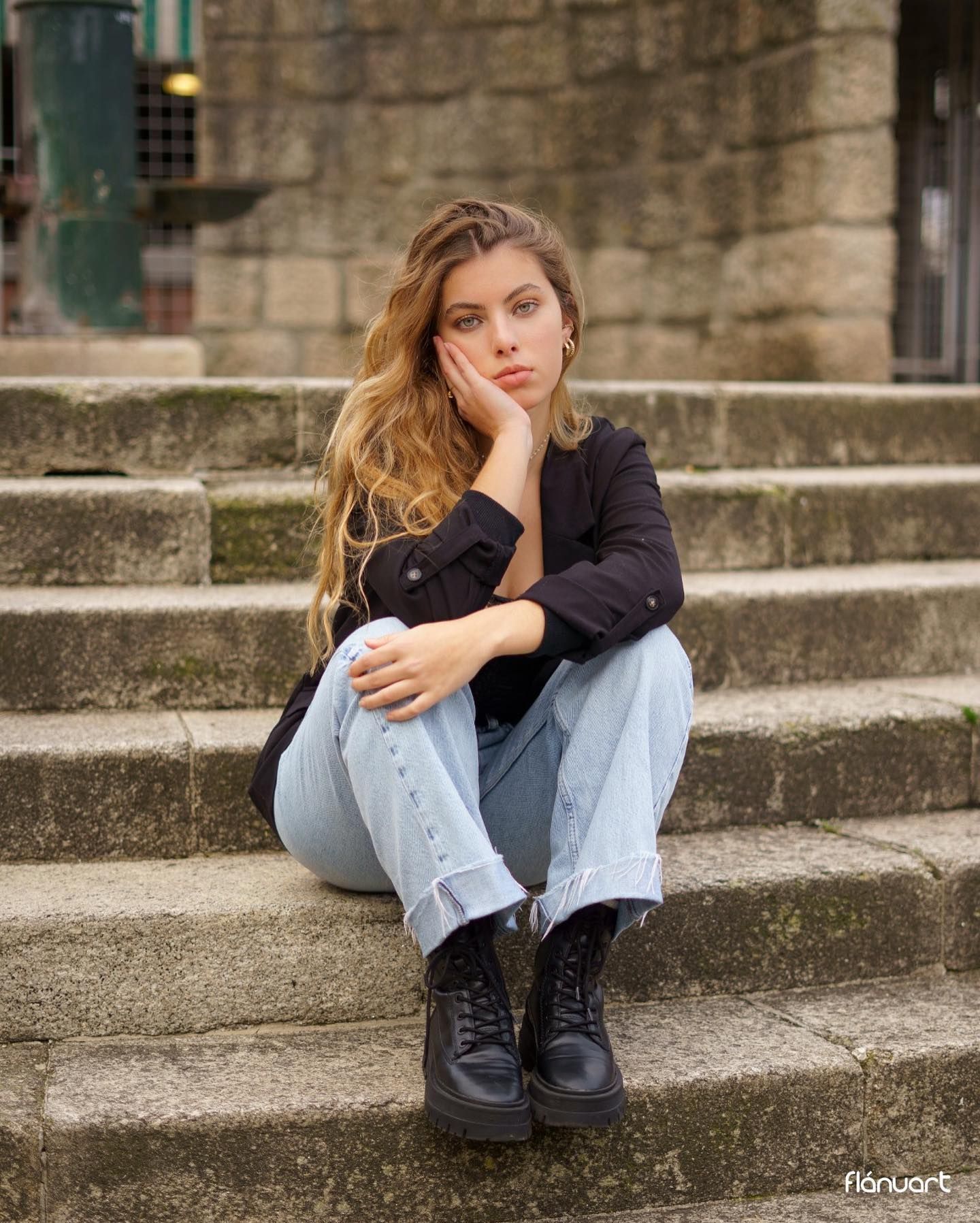 Young woman in black jacket and light jeans sitting on stone steps with combat boots.