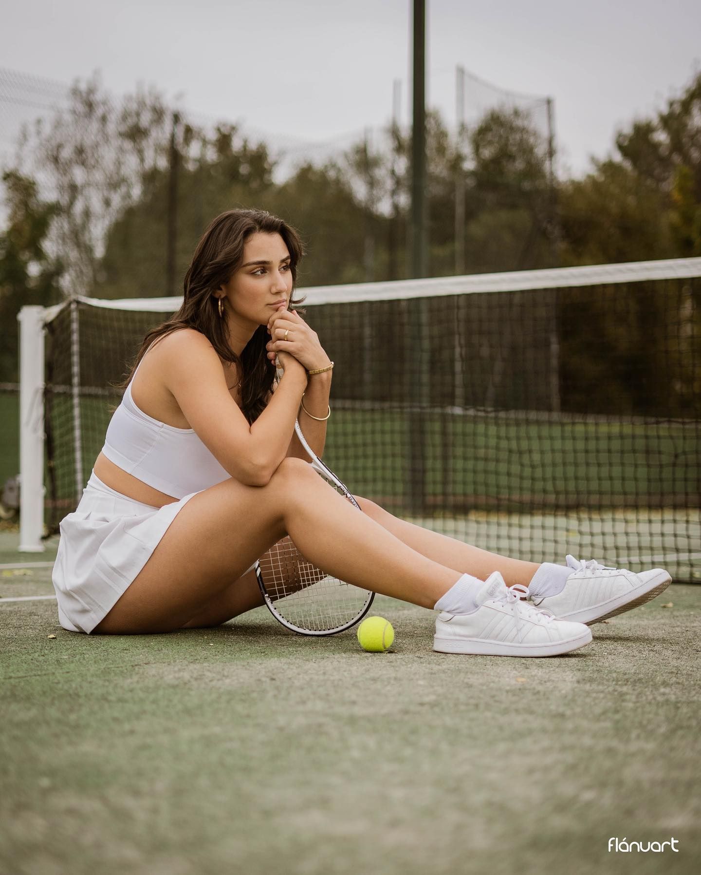 Beautiful woman in white tennis outfit sitting on a court beside a racket and tennis ball.