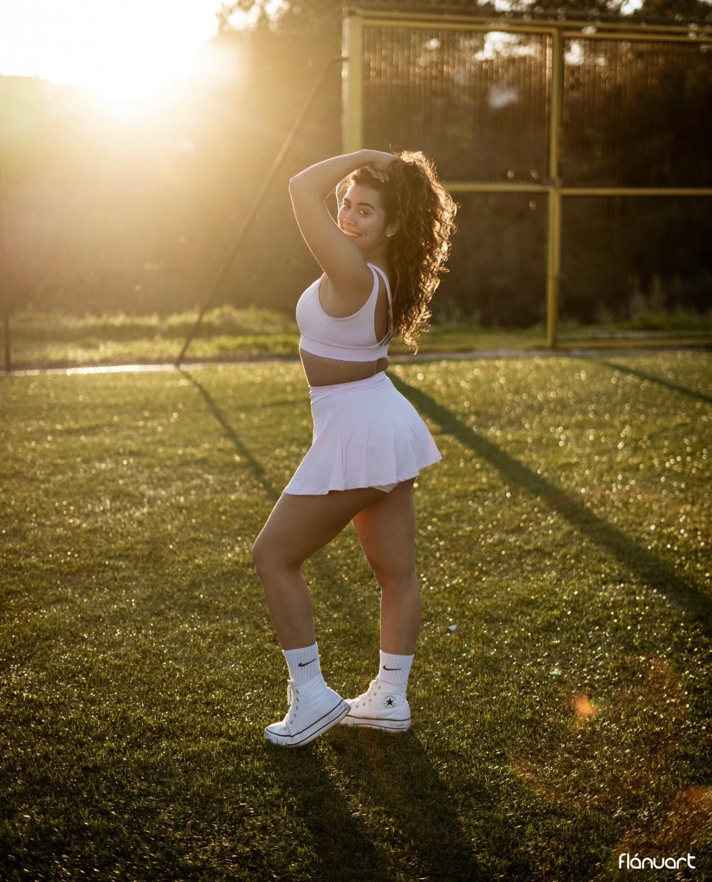Young woman in white tennis skirt and crop top posing on grass court in golden sunlight.