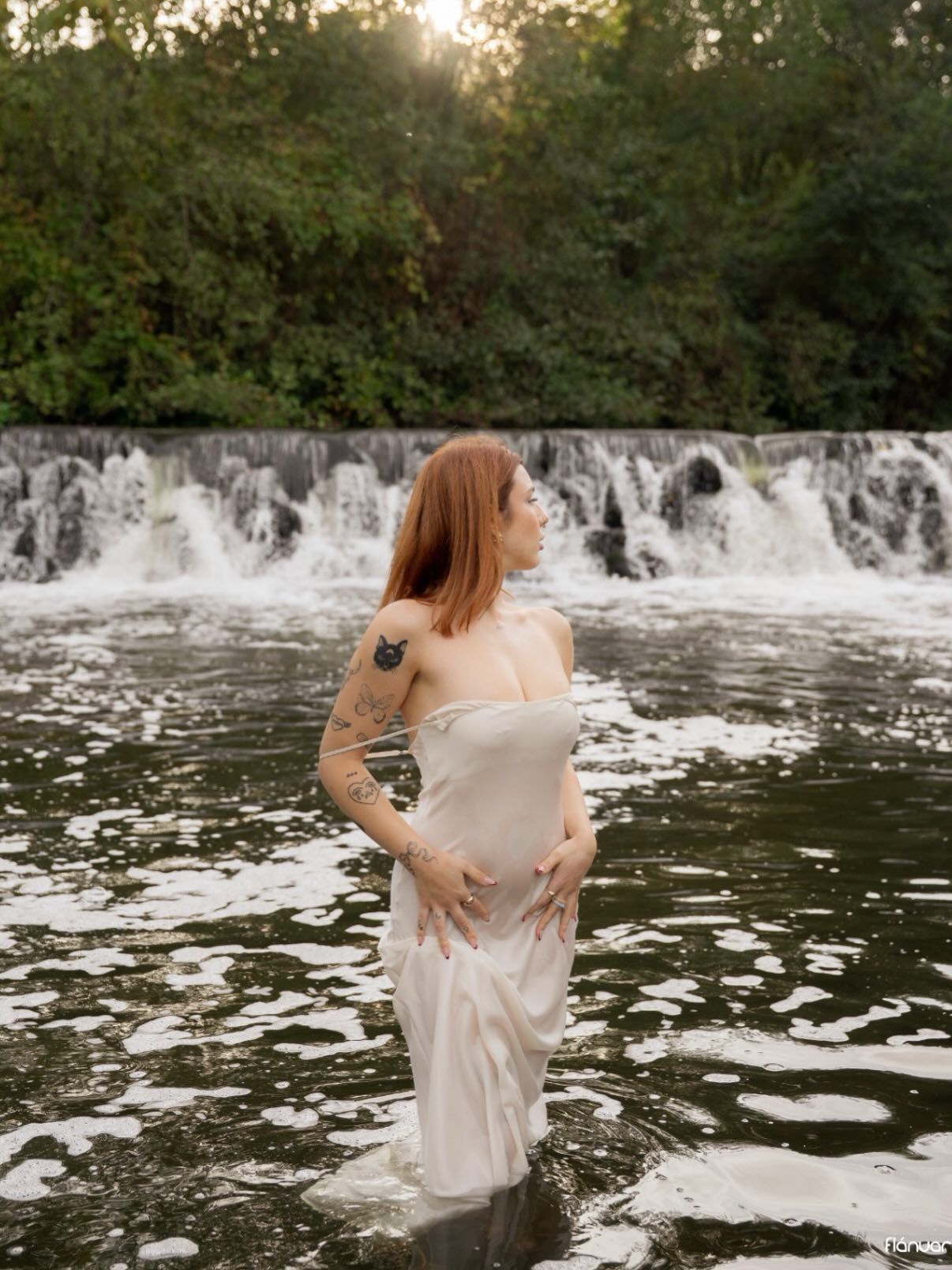 Redhead woman in sheer white dress standing in shallow river with waterfall backdrop.