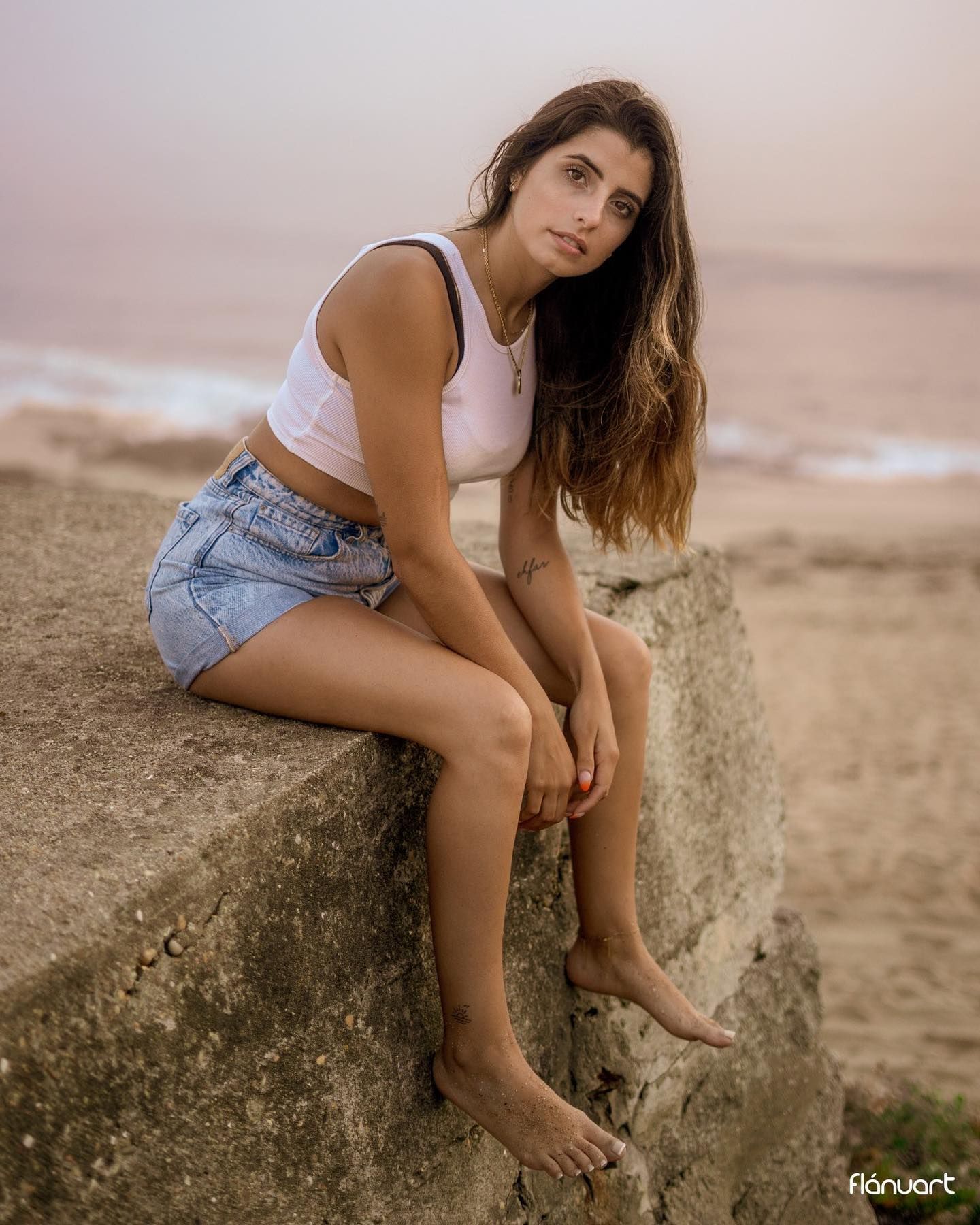 Woman in white tank top and denim shorts sitting on a concrete wall at the beach.