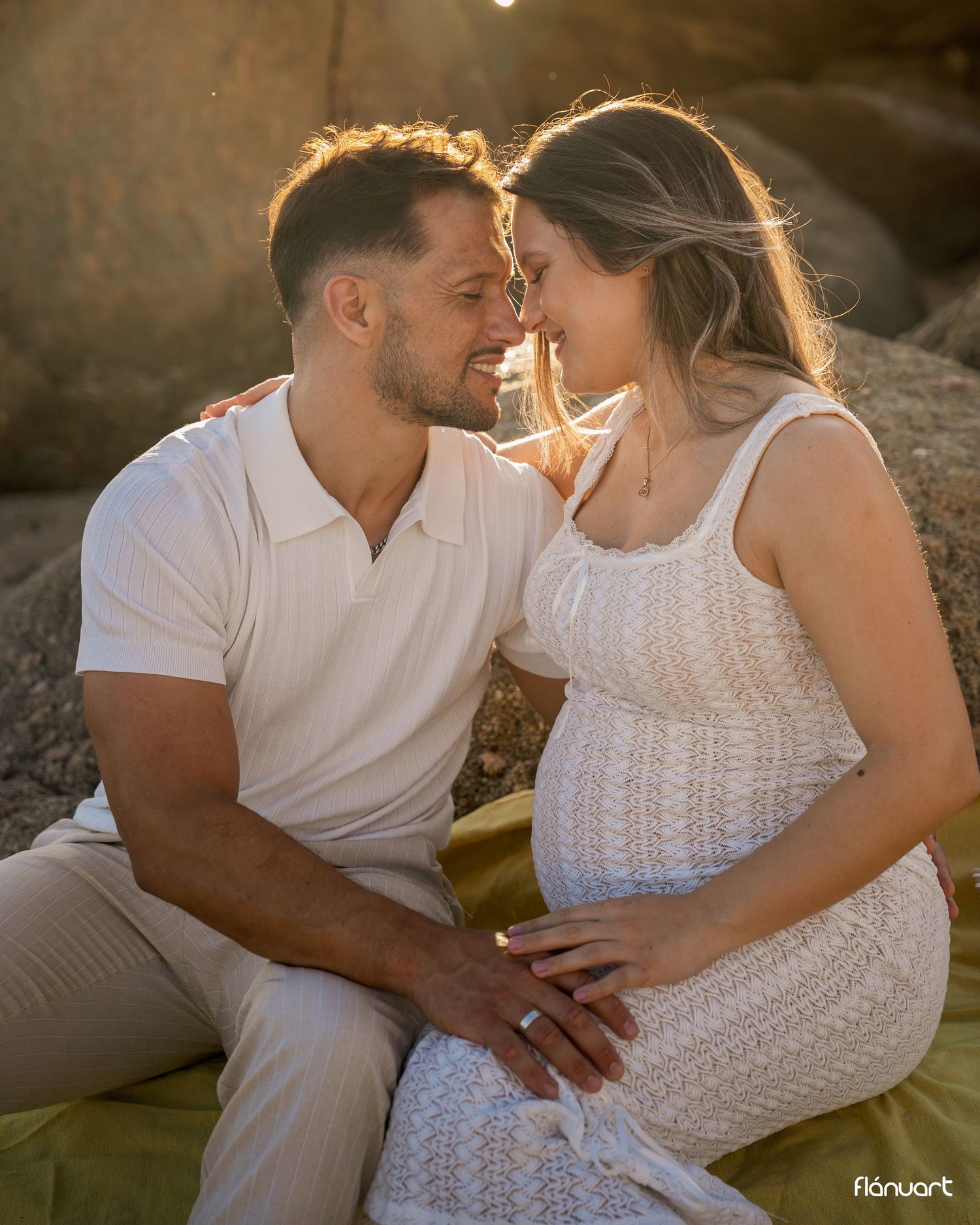 Couple sharing a romantic moment outdoors during golden hour, woman in white lace maternity dress, man in white polo.