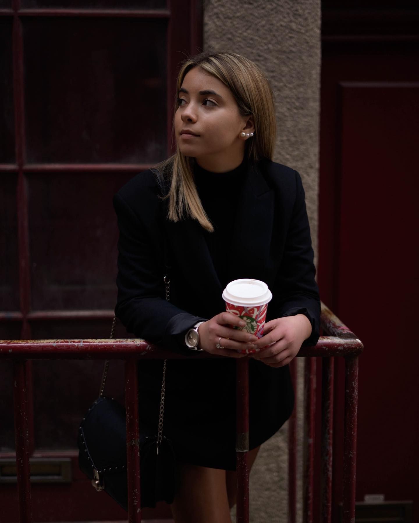 Stylish woman in black outfit holding a coffee cup while leaning on a railing outdoors.