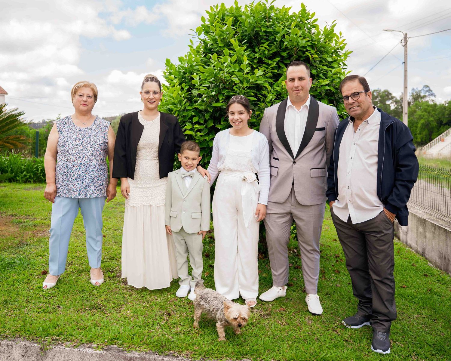 An extended family group poses outdoors with a small dog during a First Communion celebration.