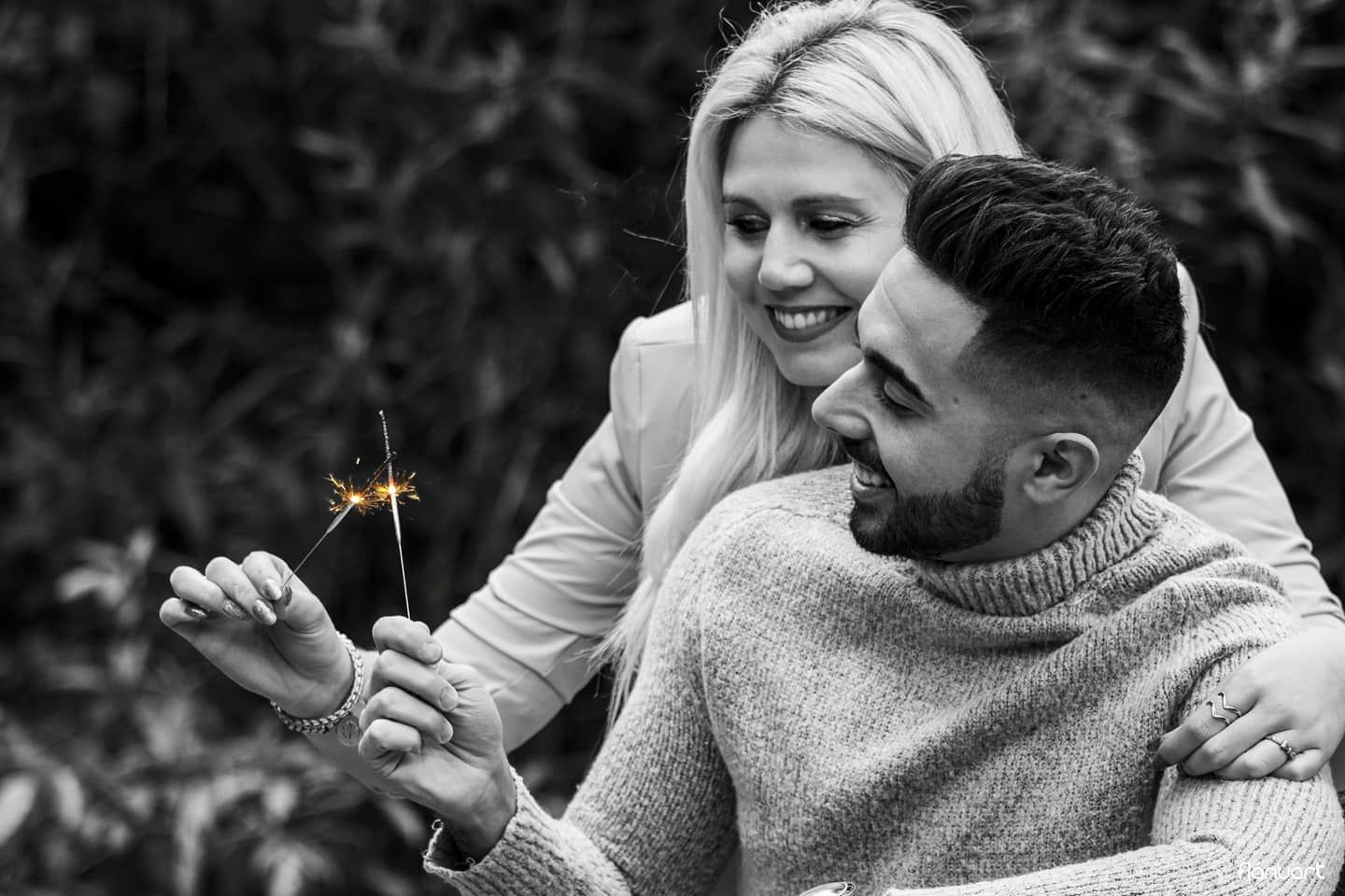 Black and white photo of couple sharing a sparkler, woman smiling behind bearded man in knit sweater.