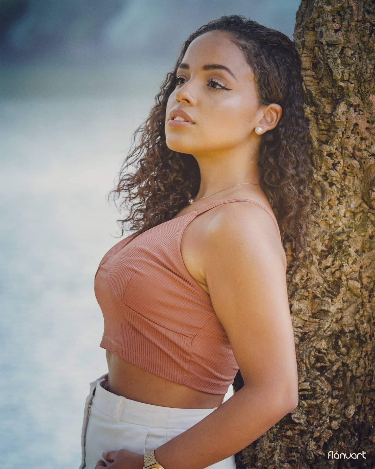 Woman with curly hair in a terracotta crop top leaning against a tree in a serene outdoor setting.