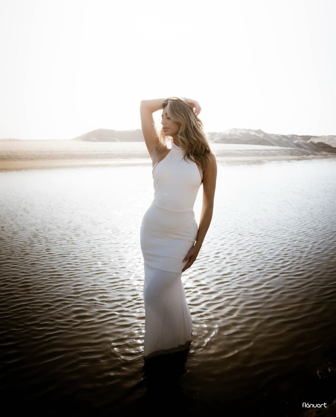 Elegant woman in white fitted gown standing in shallow water with bright sunset behind her.