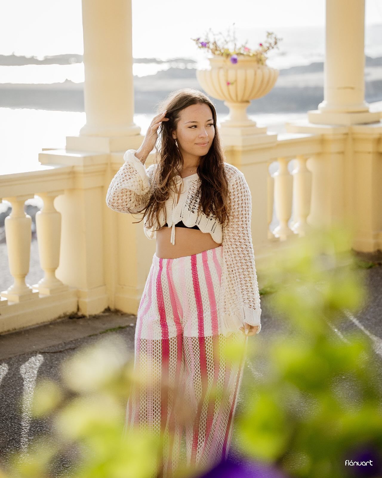 Young woman in crochet top and pink striped skirt posing on a white balustrade terrace overlooking the sea.