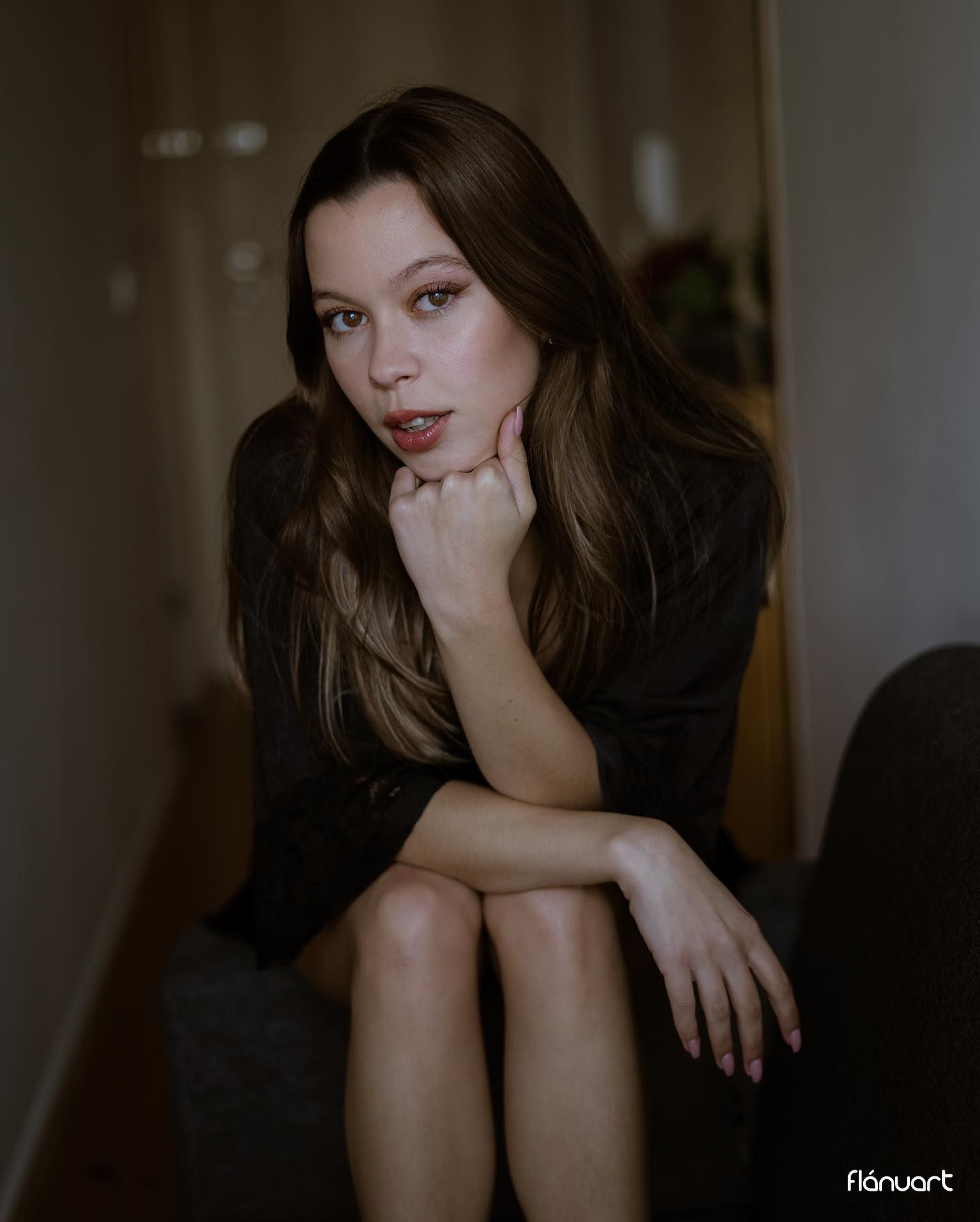 Brunette woman in dark outfit sits thoughtfully with hand under chin indoors.