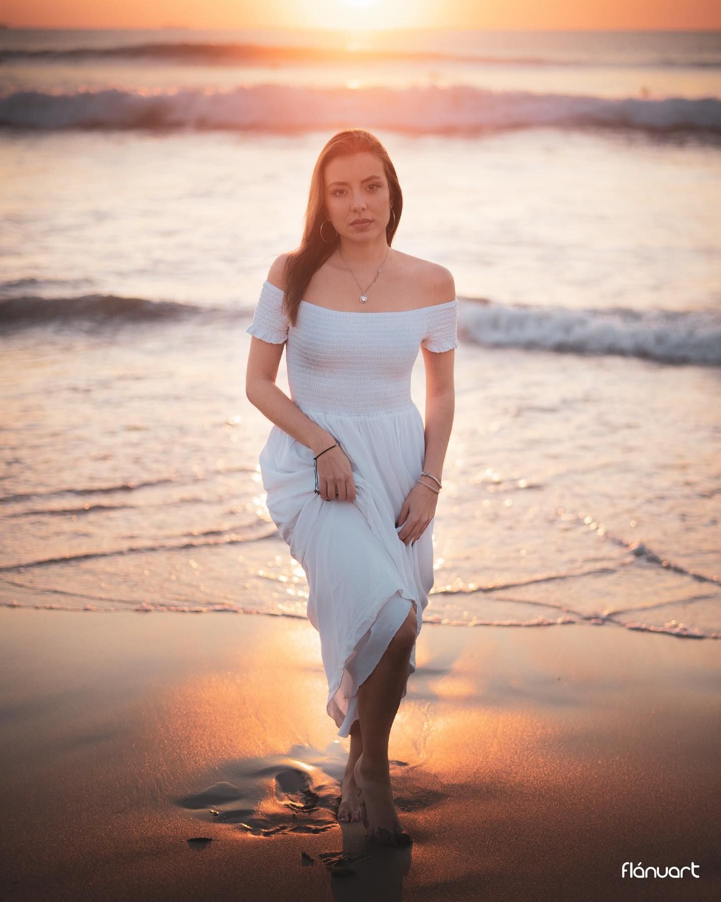 Woman in white off-shoulder dress walking on beach at sunset.