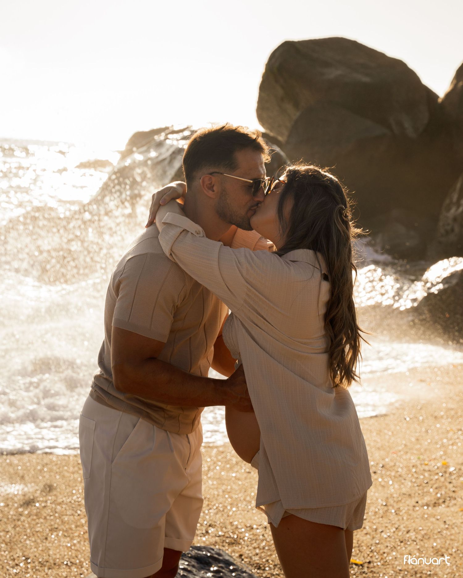 Couple kissing romantically on a sunny beach with crashing waves and large rocks in the background.