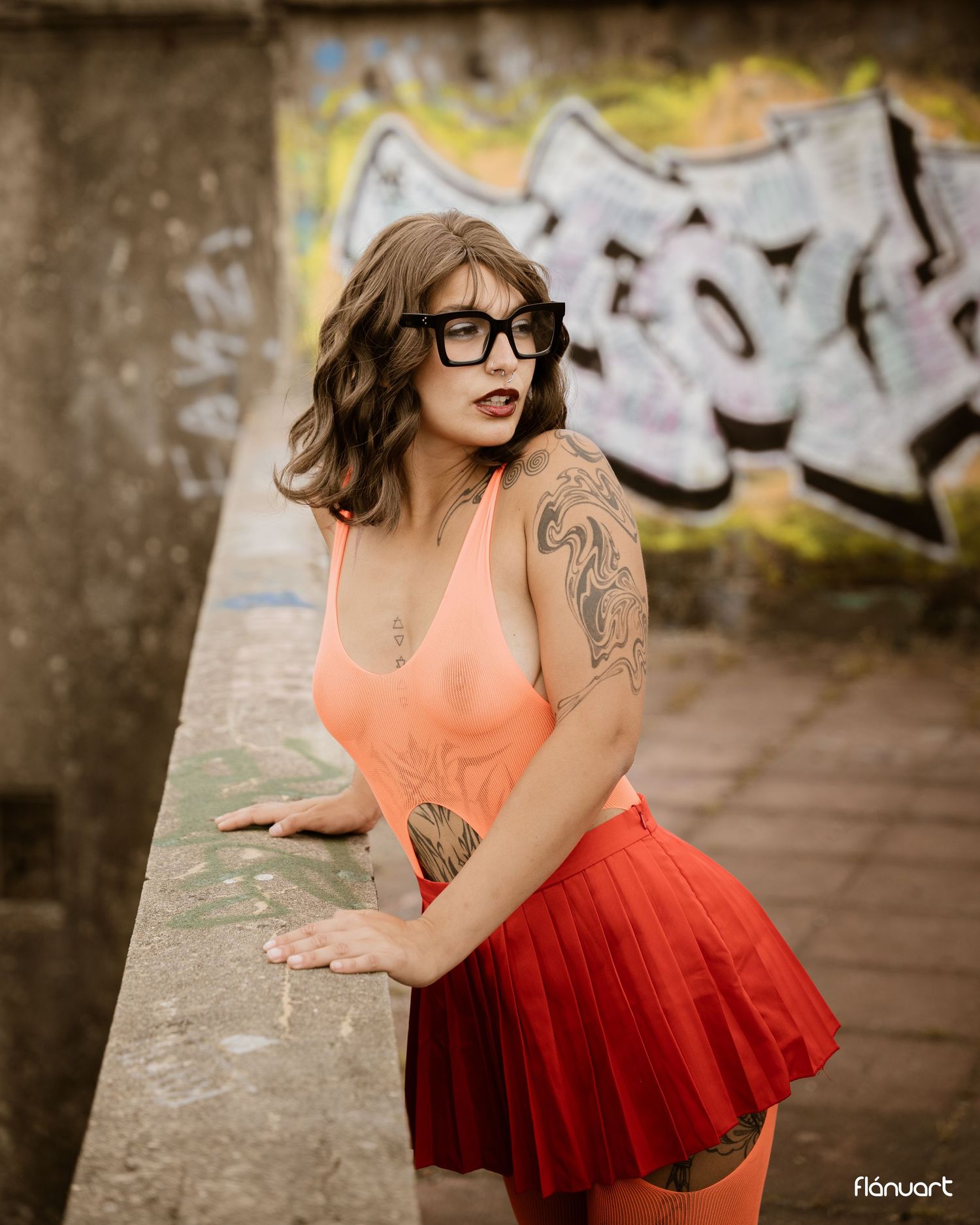 Young woman with tattoos in orange bodysuit and red skirt poses near graffiti wall wearing black glasses.