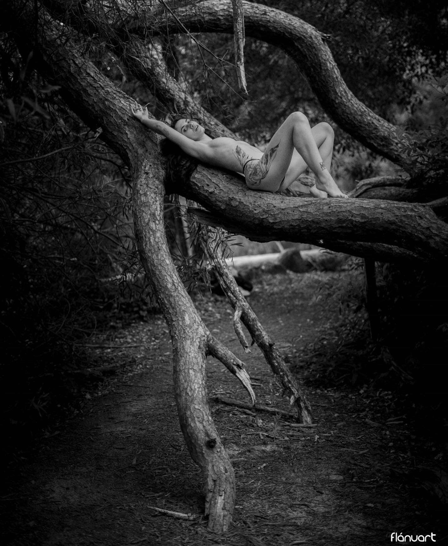 Black and white artistic photo of woman resting on twisted tree branches in forest.