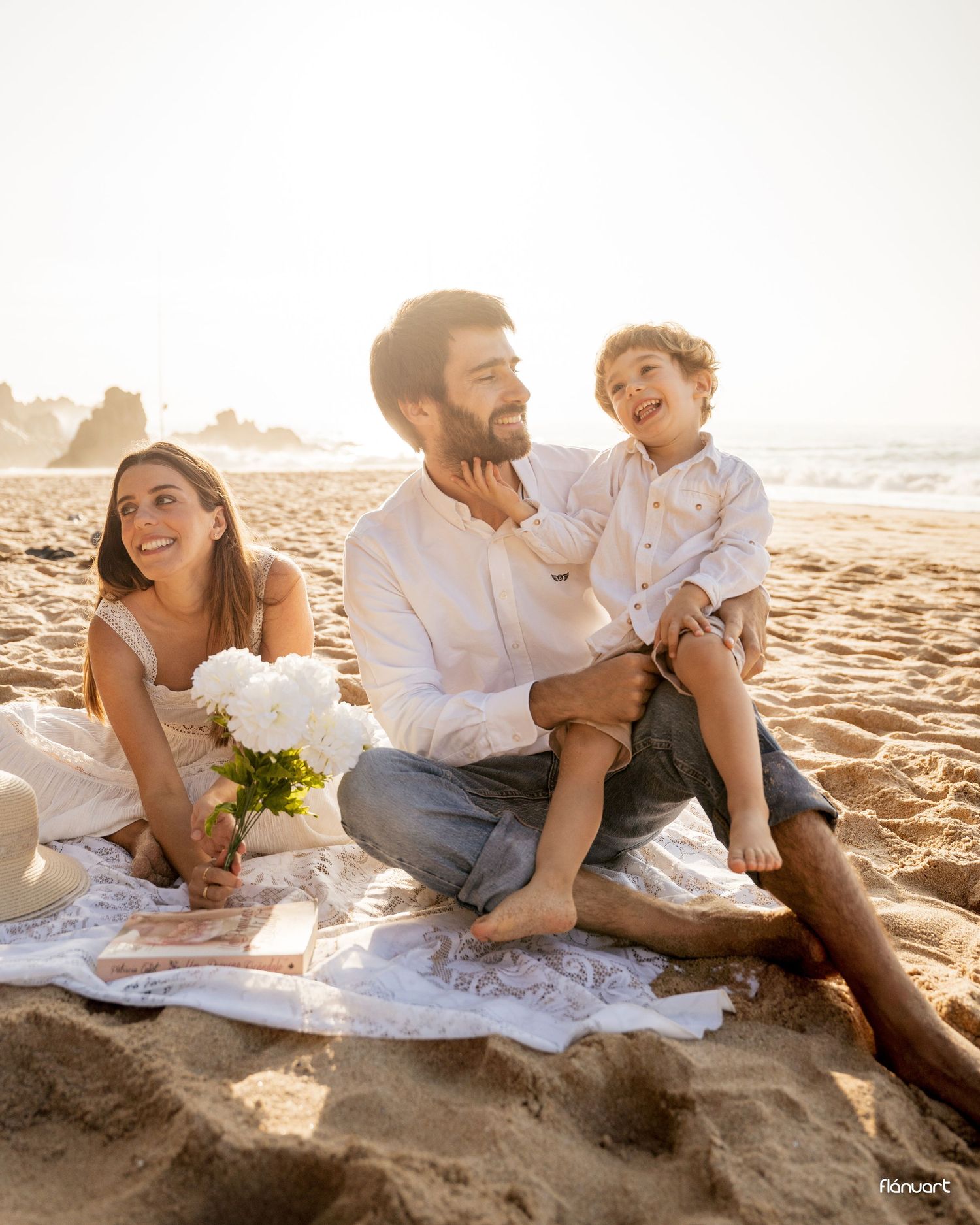 Loving family sitting on sandy beach at sunset, father holding young son while mother smiles.