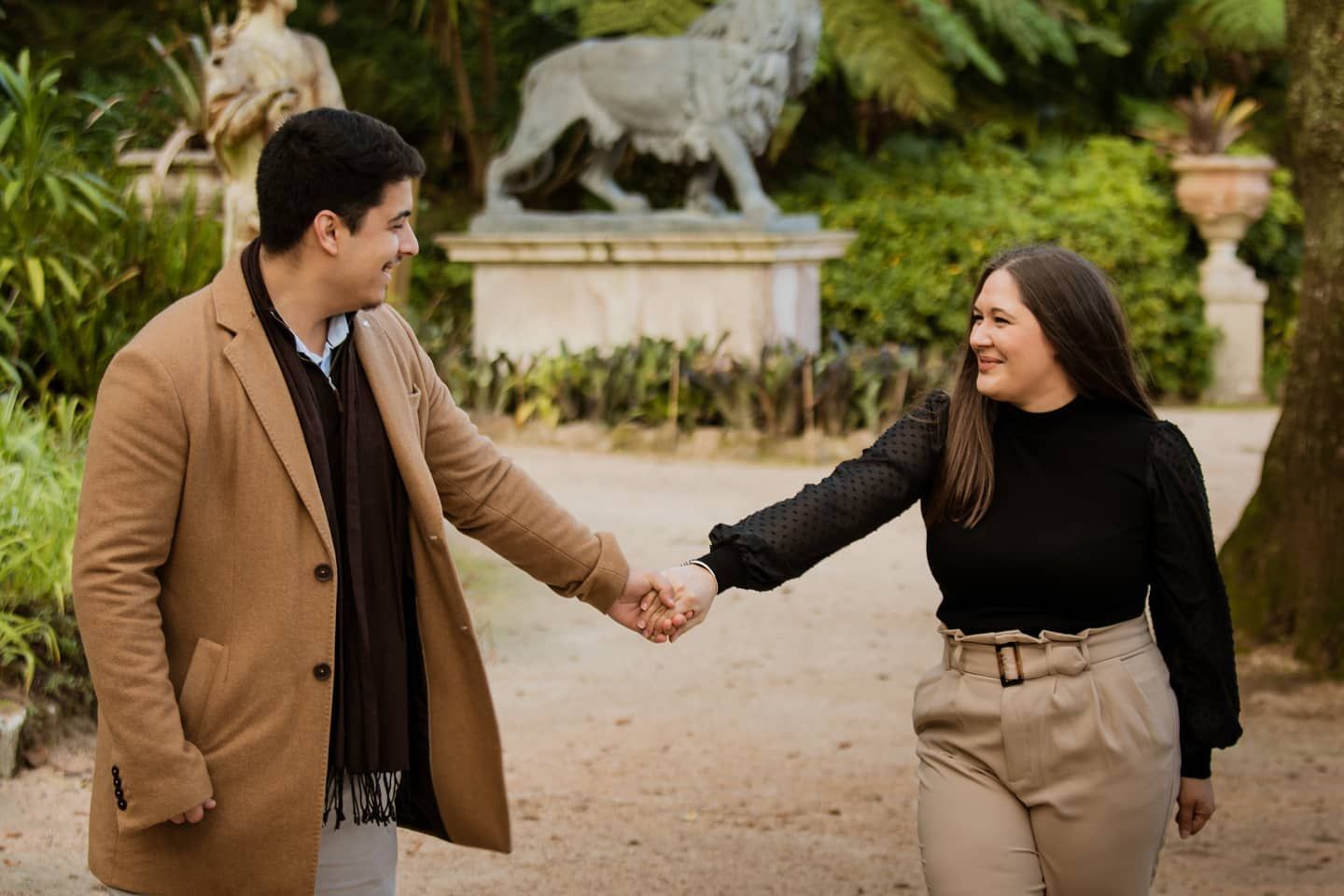 Smiling couple holding hands in a garden park, man in camel coat, woman in black top.