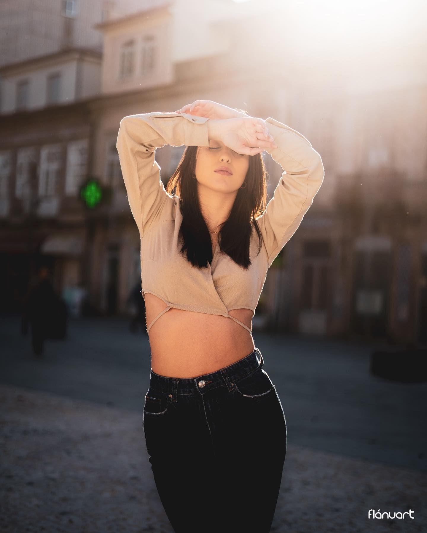 Dark-haired woman in a beige wrap crop top and black jeans posing in a sunlit city square.