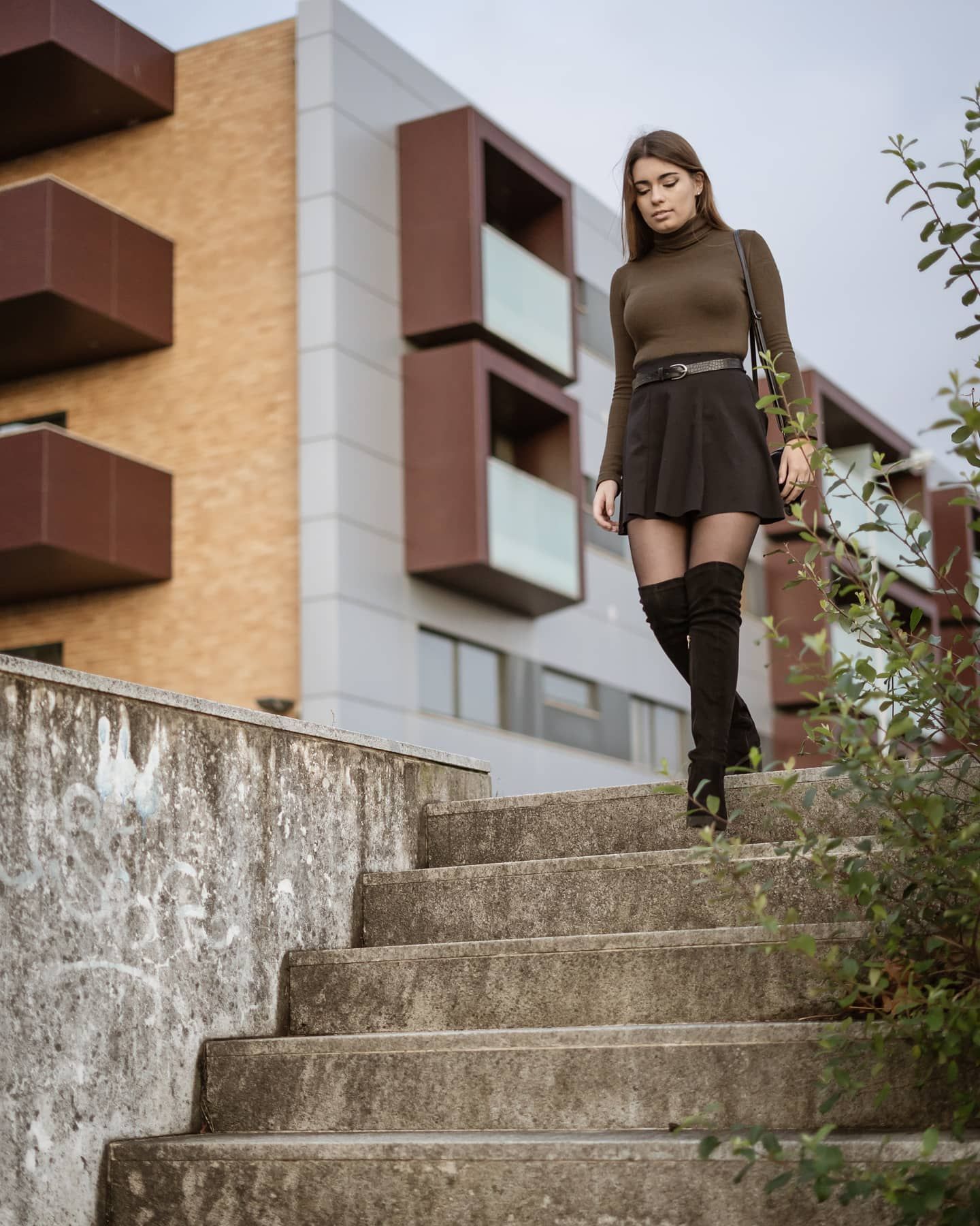 Stylish woman in brown turtleneck, mini skirt, and thigh-high boots descending outdoor urban staircase.