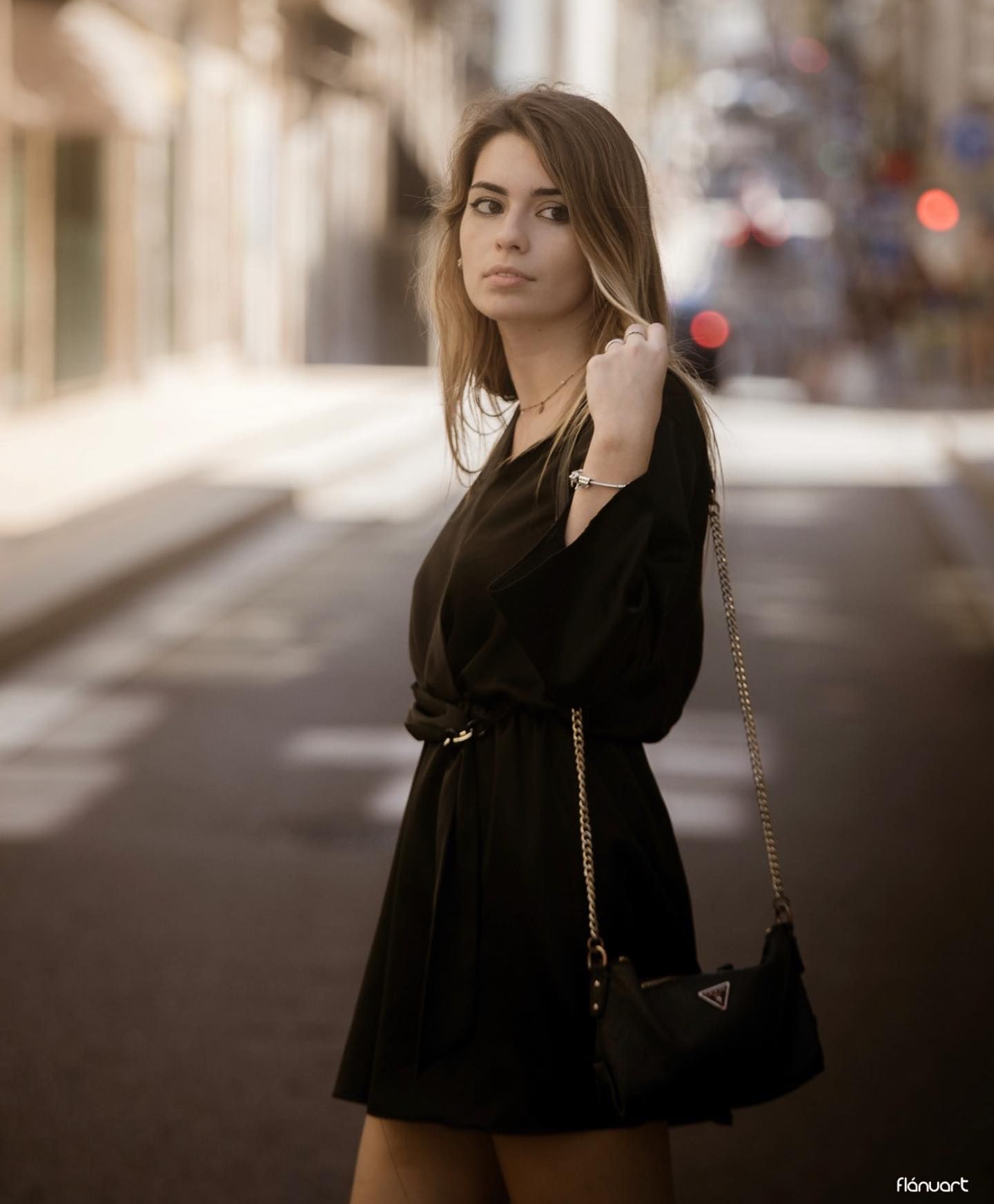 Young woman in black wrap dress with chain bag poses on a blurred city street.
